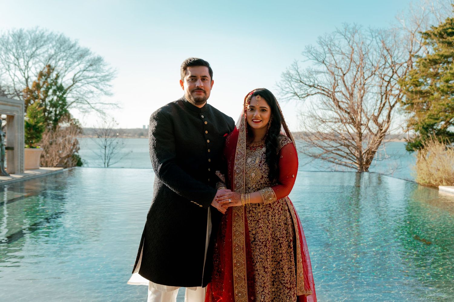 Couple in formal attire posing near a waterfront with blue sky and trees in background.