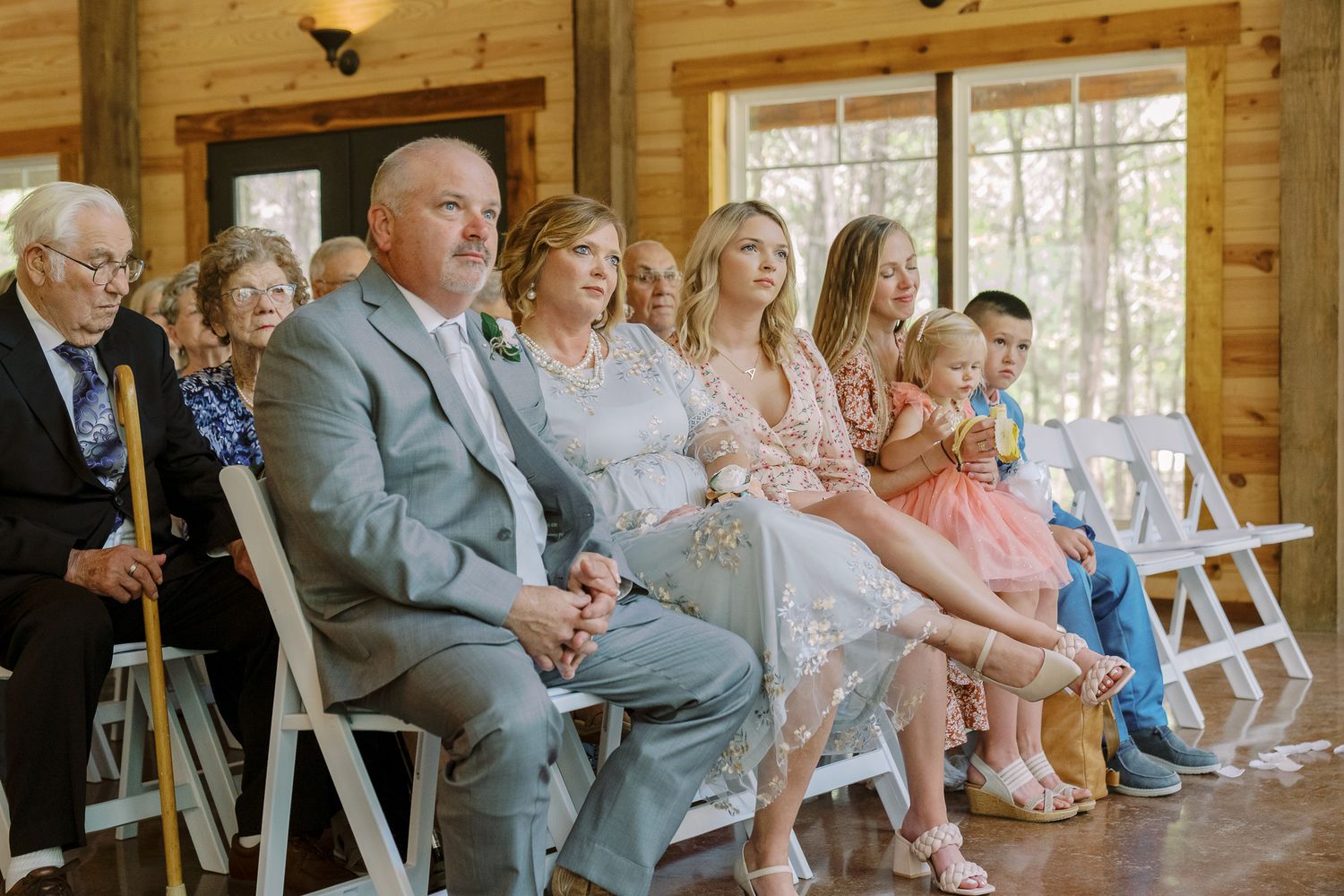 Wedding guests seated in wooden chairs watch a ceremony in a sunlit rustic venue with natural decor.
