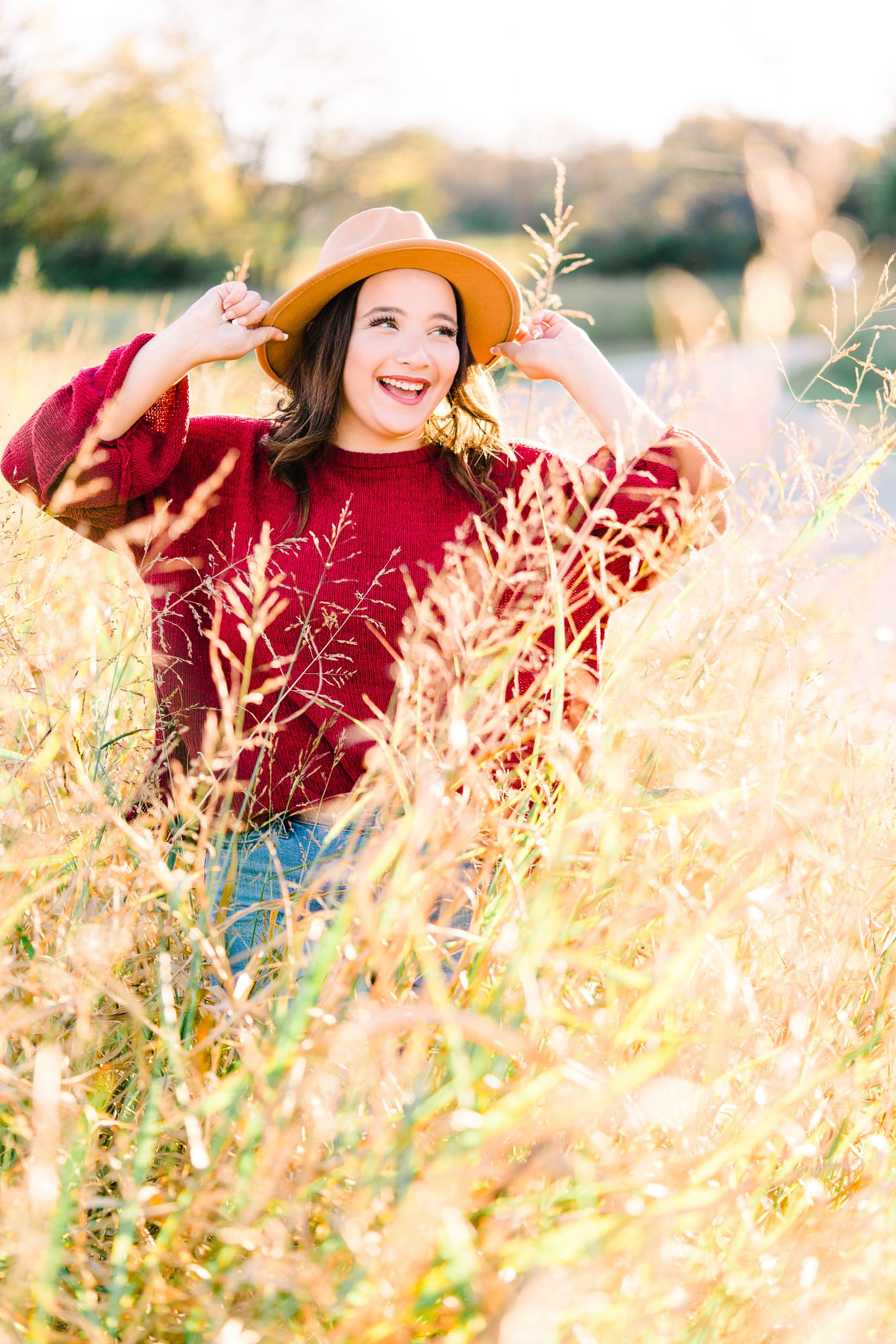 Person in red sweater and straw hat poses joyfully in golden field during sunset.