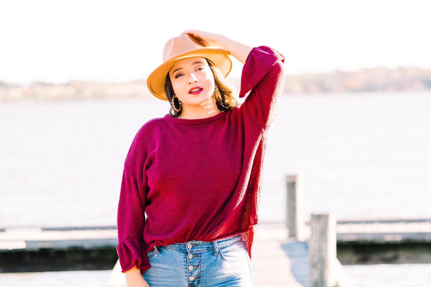 Fashion model in burgundy sweater and tan hat poses casually on wooden dock by waterfront at sunset.