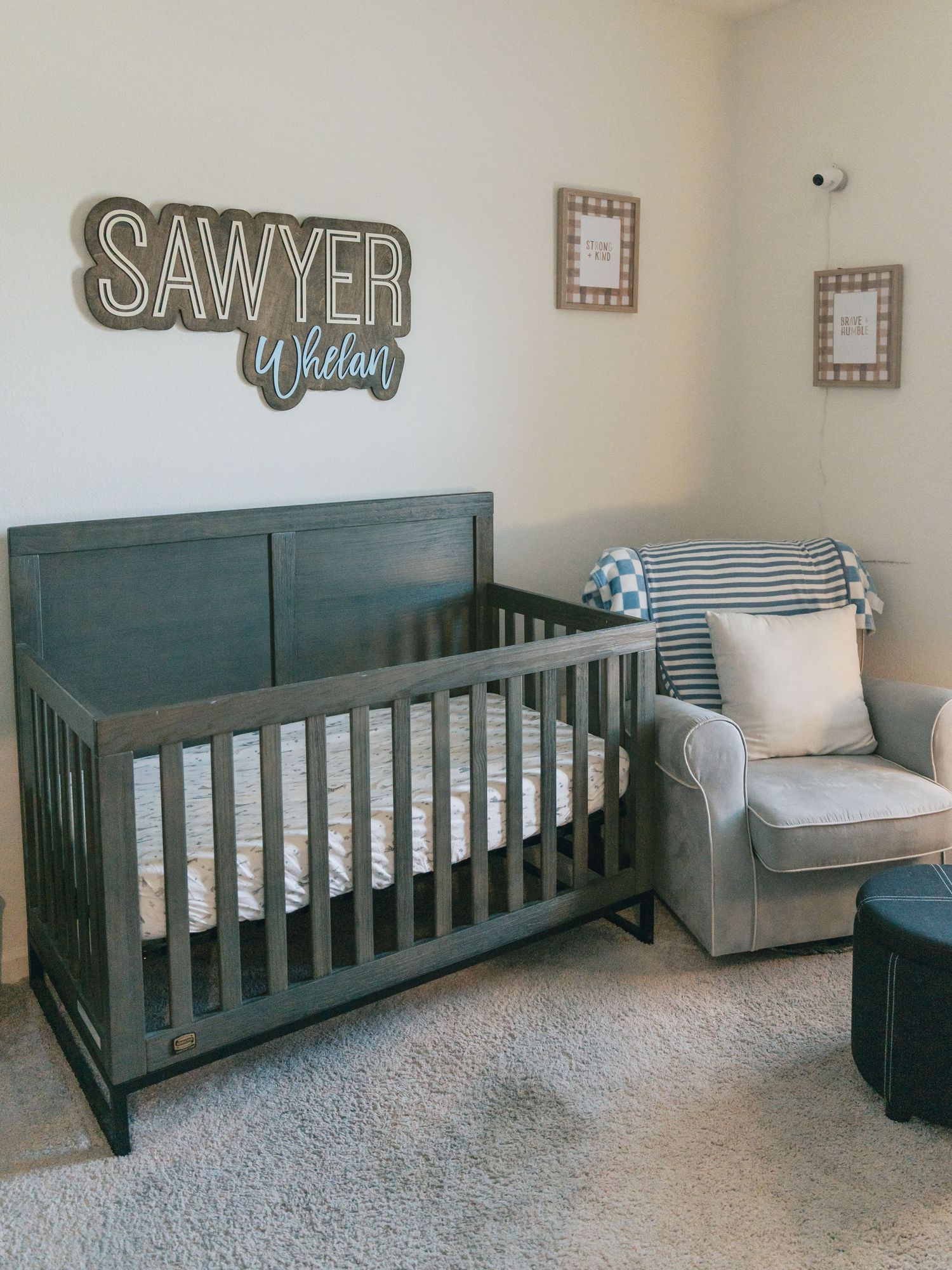 Nursery room setup with dark teal crib, gray armchair, and 'Sawyer' name sign mounted on neutral walls.