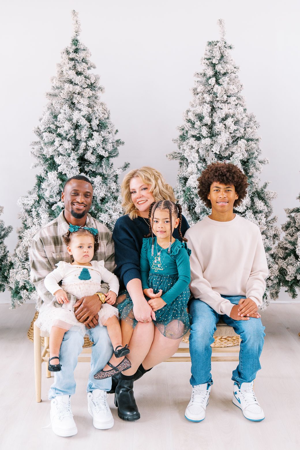 Family in coordinating teal and white outfits sits together against snowy Christmas trees for holiday photos.