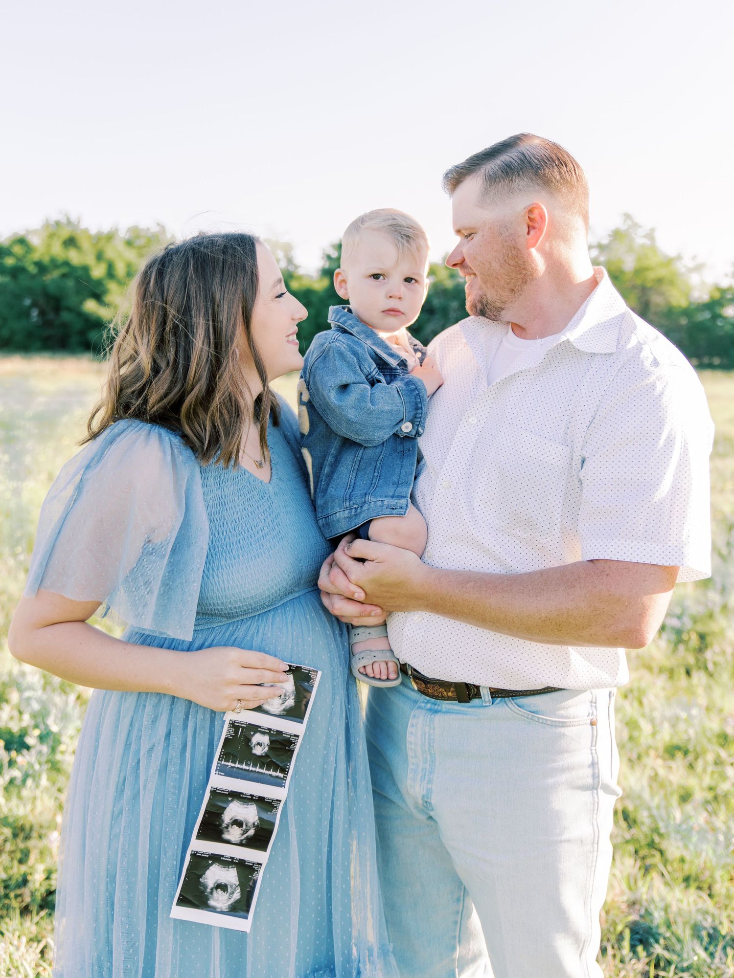 Family sharing tender moments while holding ultrasound photos in a sunlit field.