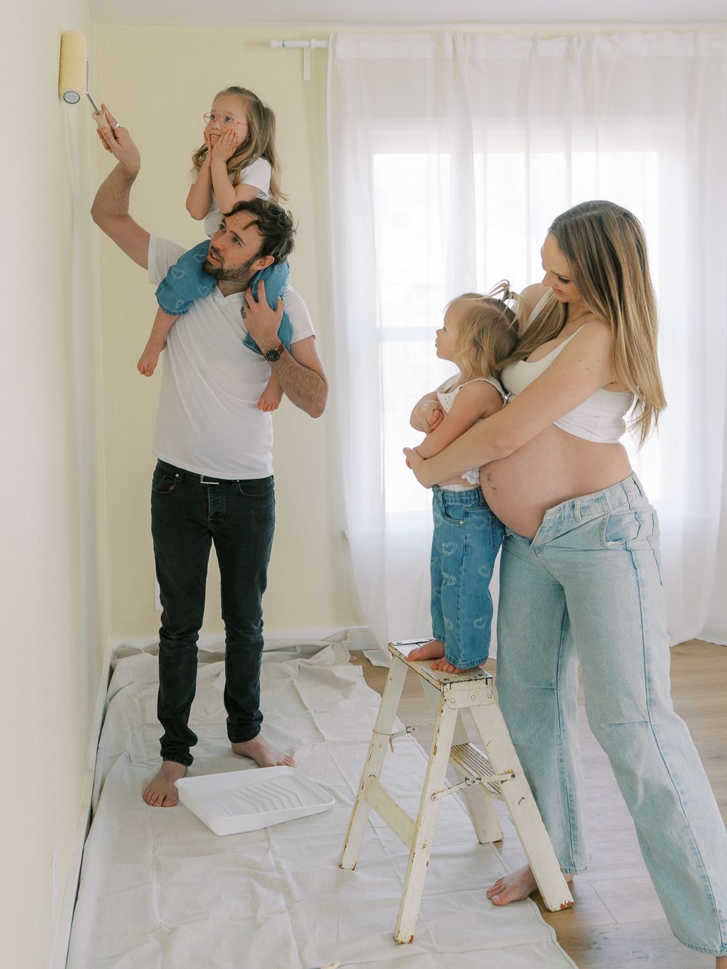 Family painting interior walls together with child on shoulders and toddler on stepladder in bright white room.