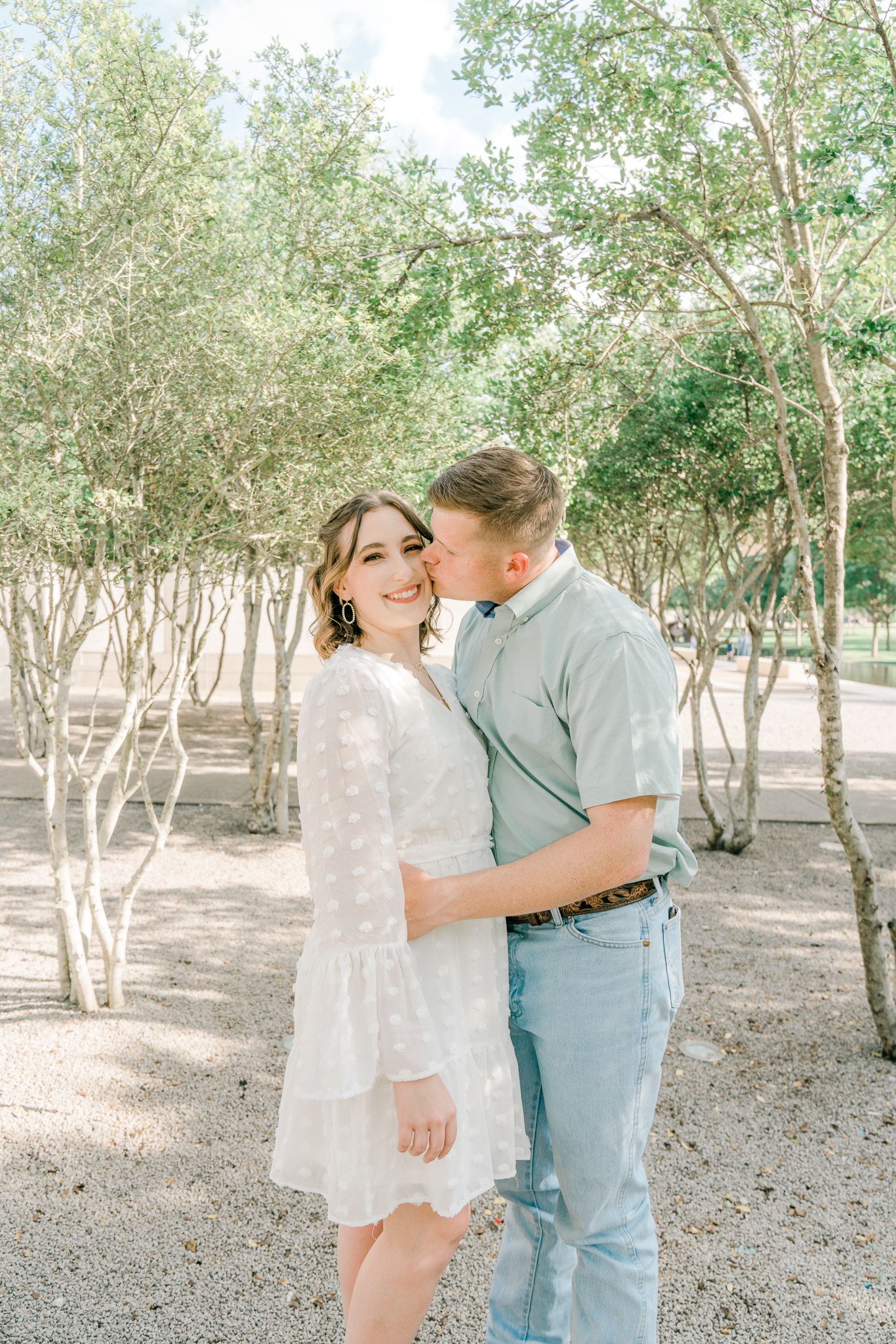 A couple embraces tenderly in a grove of trees during a bright sunny day.