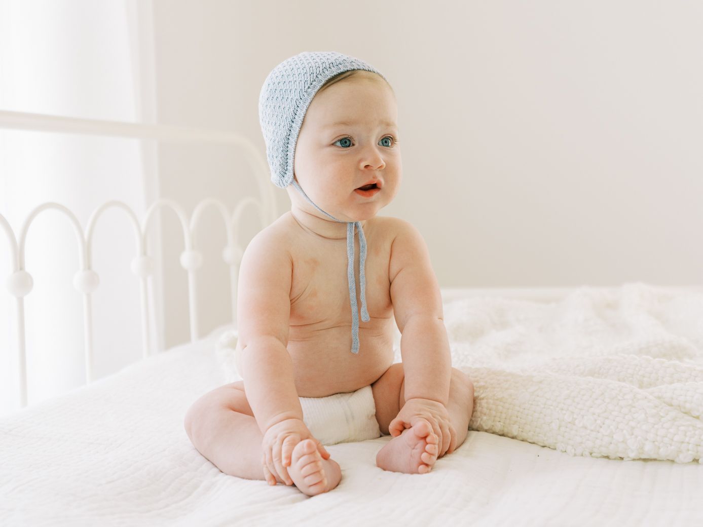 Baby in white onesie crawls on white bedding in bright, airy room.