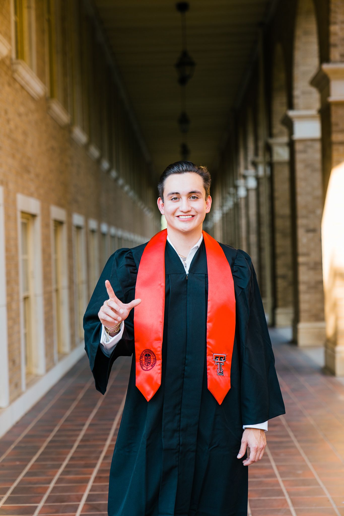 Graduate in black cap and gown with red stole smiling and gesturing in an ornate hallway.