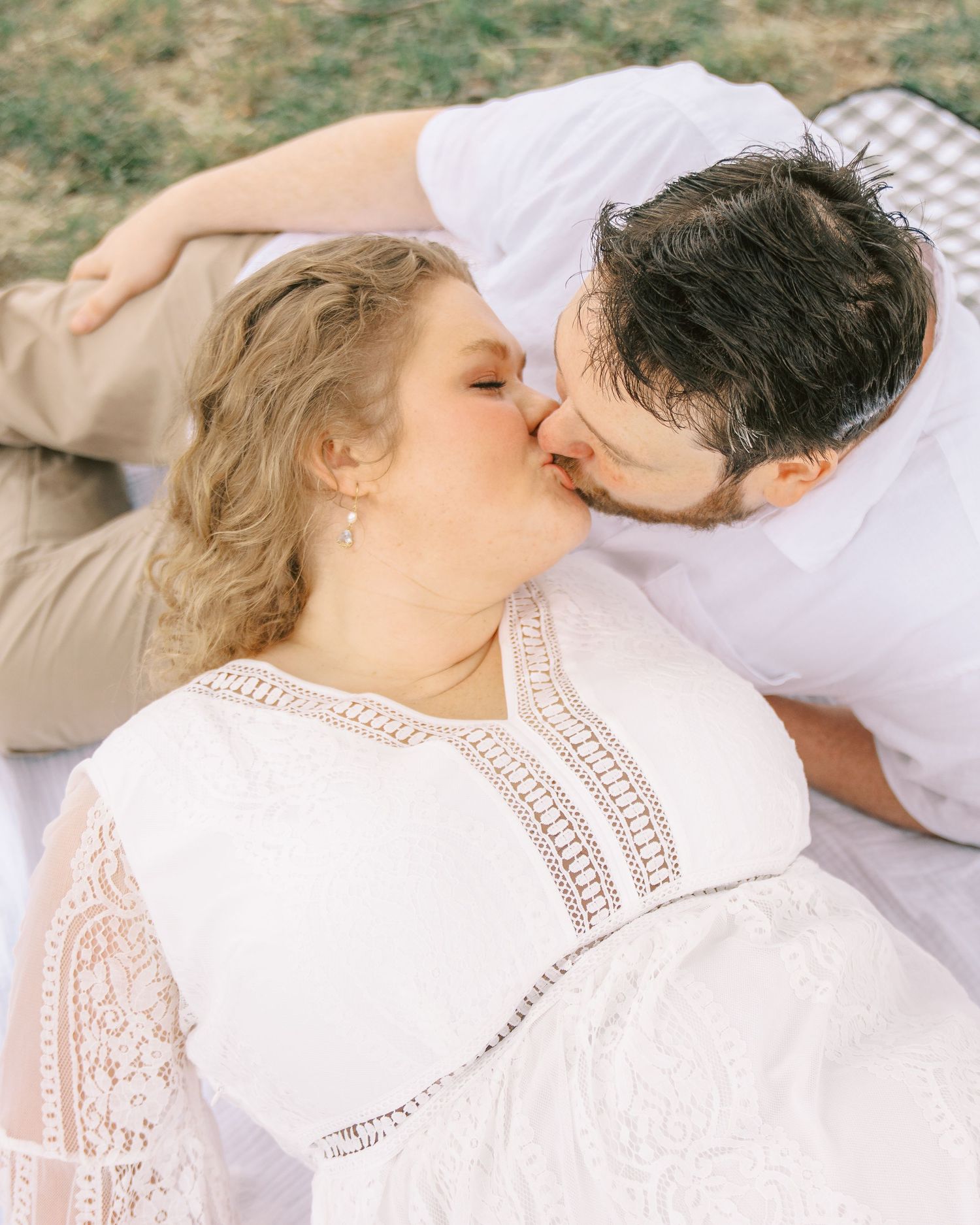 A couple shares a romantic kiss while lying on a blanket outdoors during an engagement photo session.