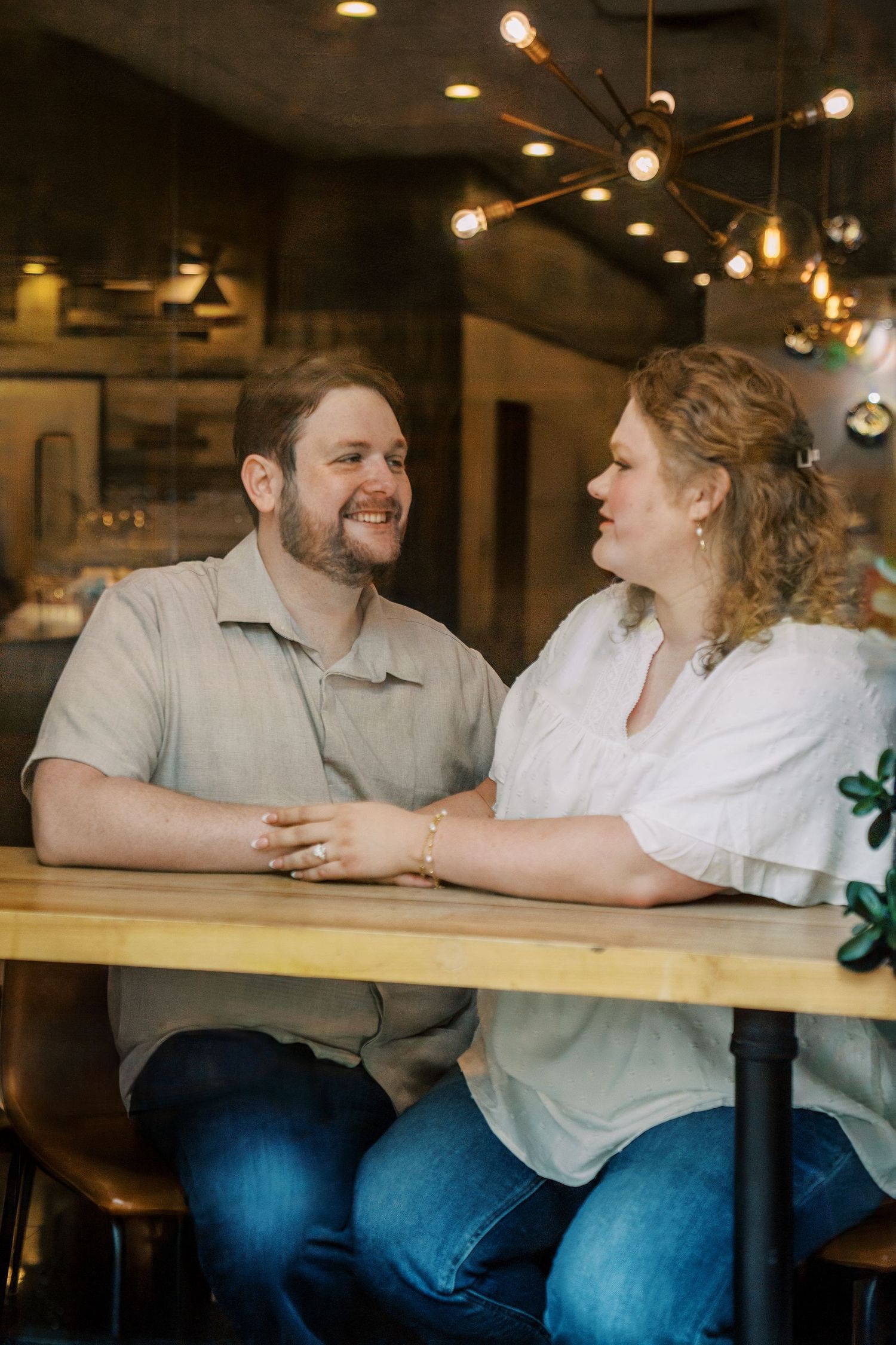 Happy engaged couple holding hands and laughing together at a wooden table inside a cozy cafe with warm lighting.