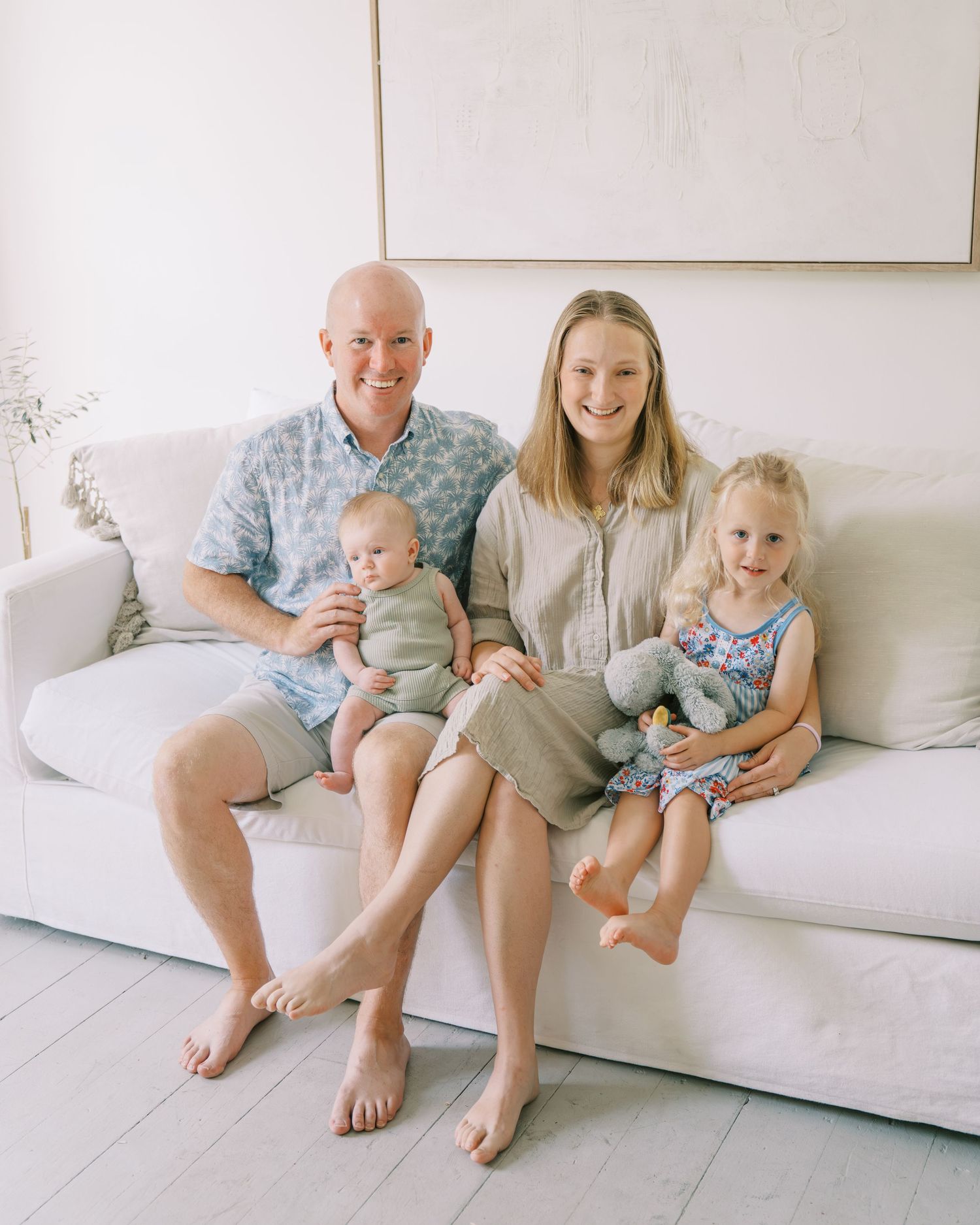 Happy family of four posing on a white couch, parents holding two young children in a bright, modern living room.