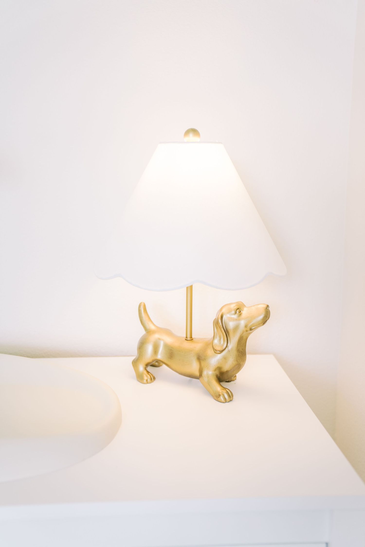 Gold dachshund dog figurine table lamp with white shade on a white nightstand.
