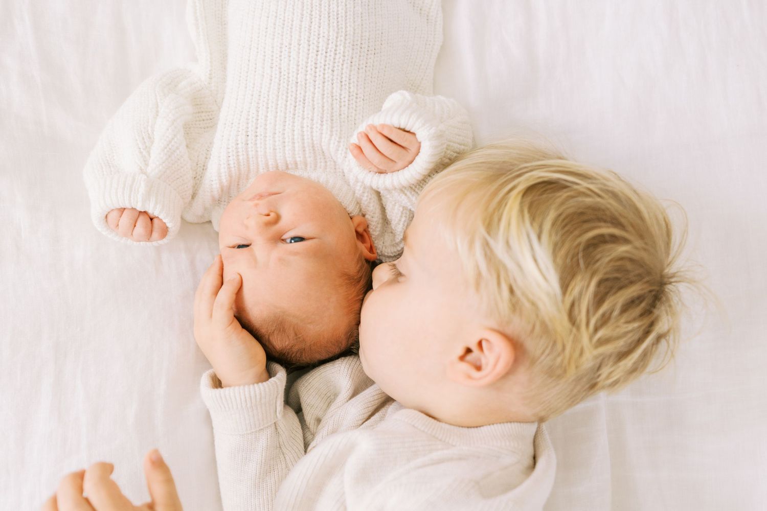 Blonde toddler boy gently kissing a sleeping newborn baby sibling while lying on a white bed.