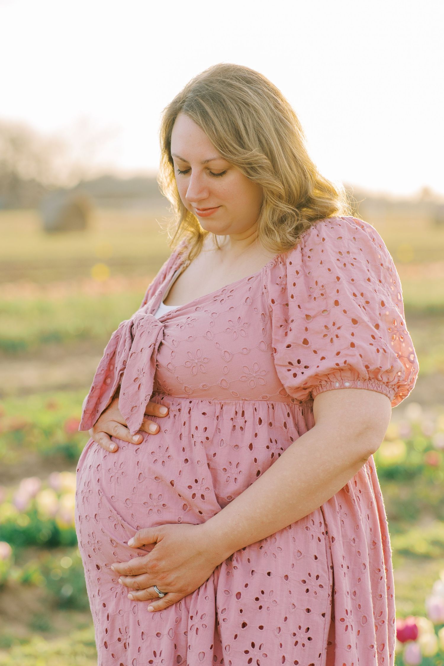 Pregnant woman in pink eyelet dress cradling baby bump in sunlit flower field during golden hour.