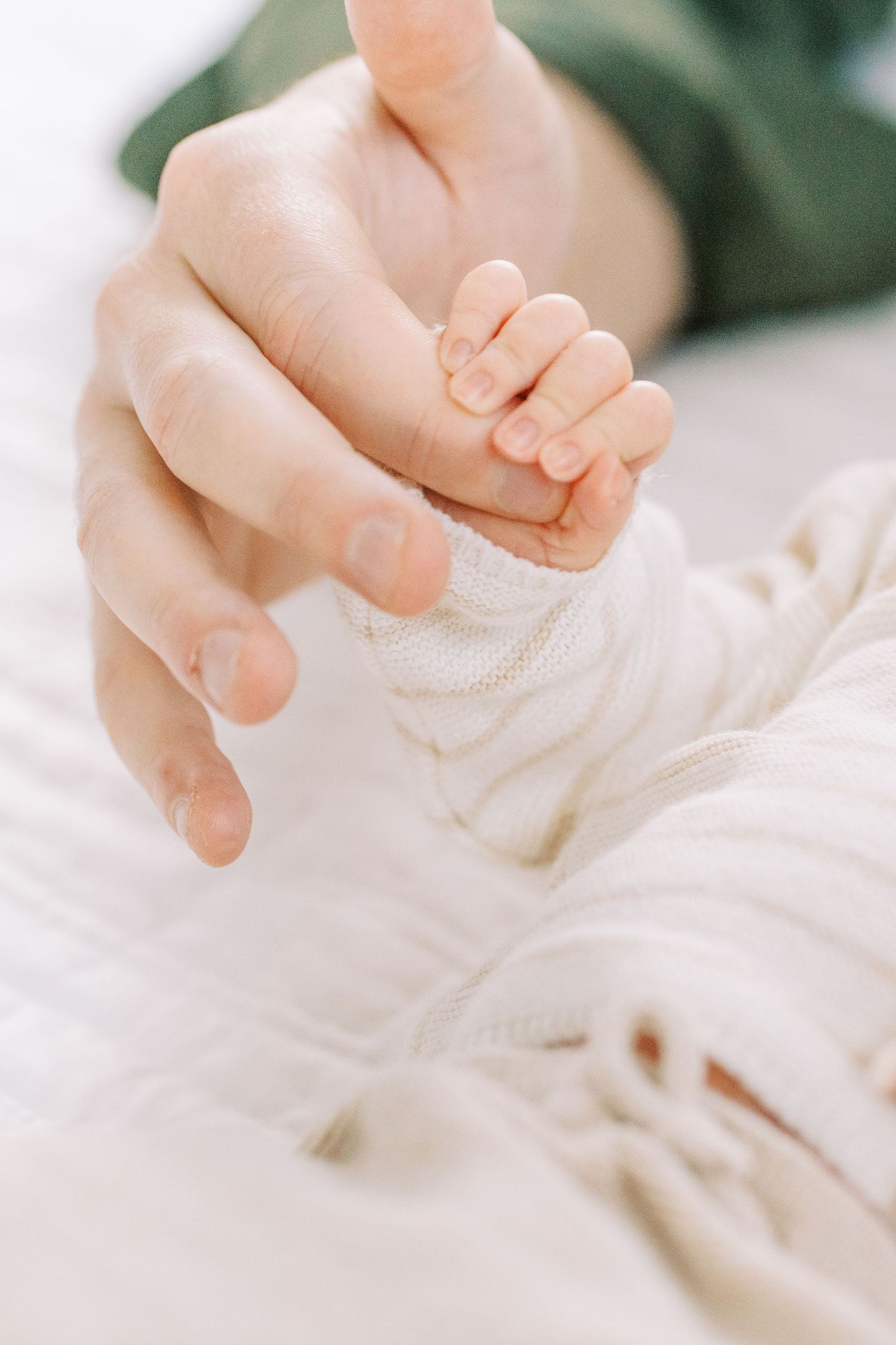 A tiny baby hand gently grasps an adult finger against soft white fabric in a tender moment of connection.