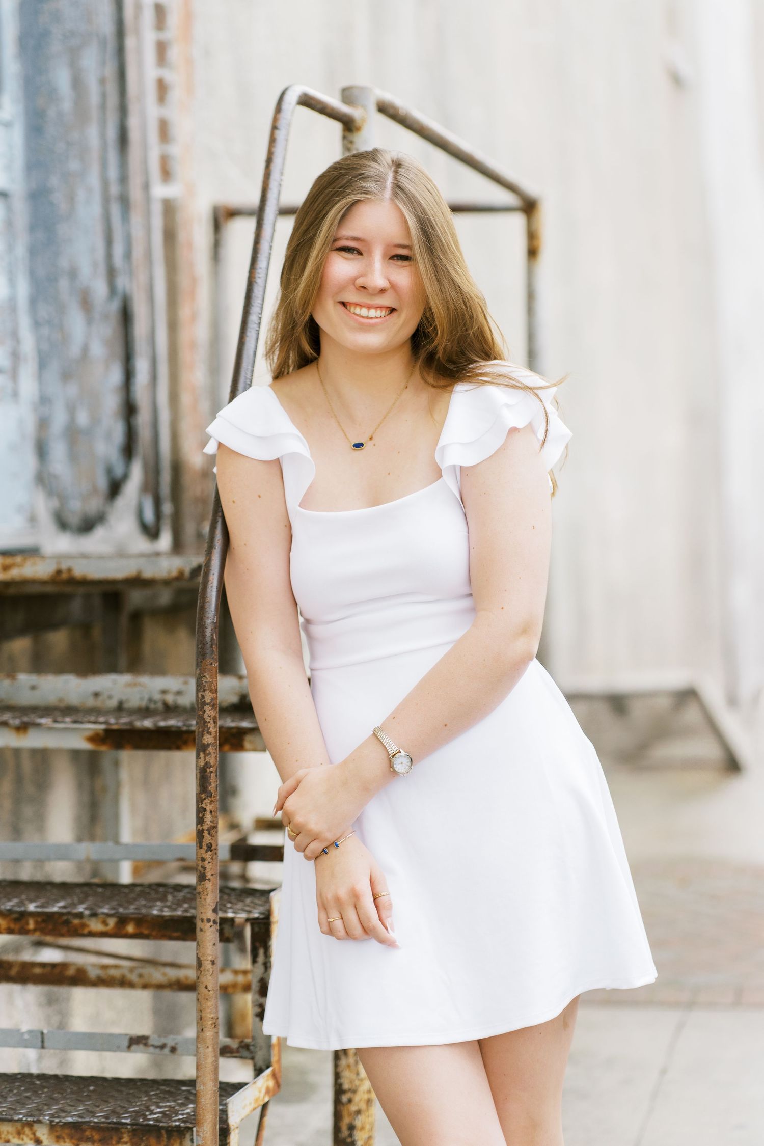 A person in a white summer dress stands smiling on rustic metal stairs against a light-colored wall.