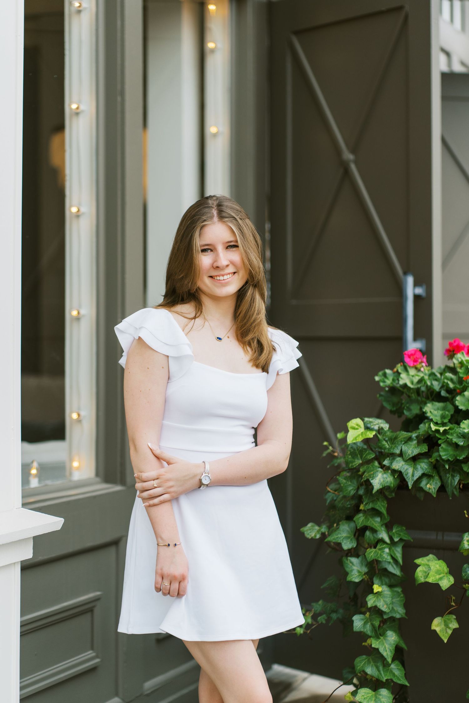 A person in a white summer dress stands on a porch with string lights and green foliage in the background.