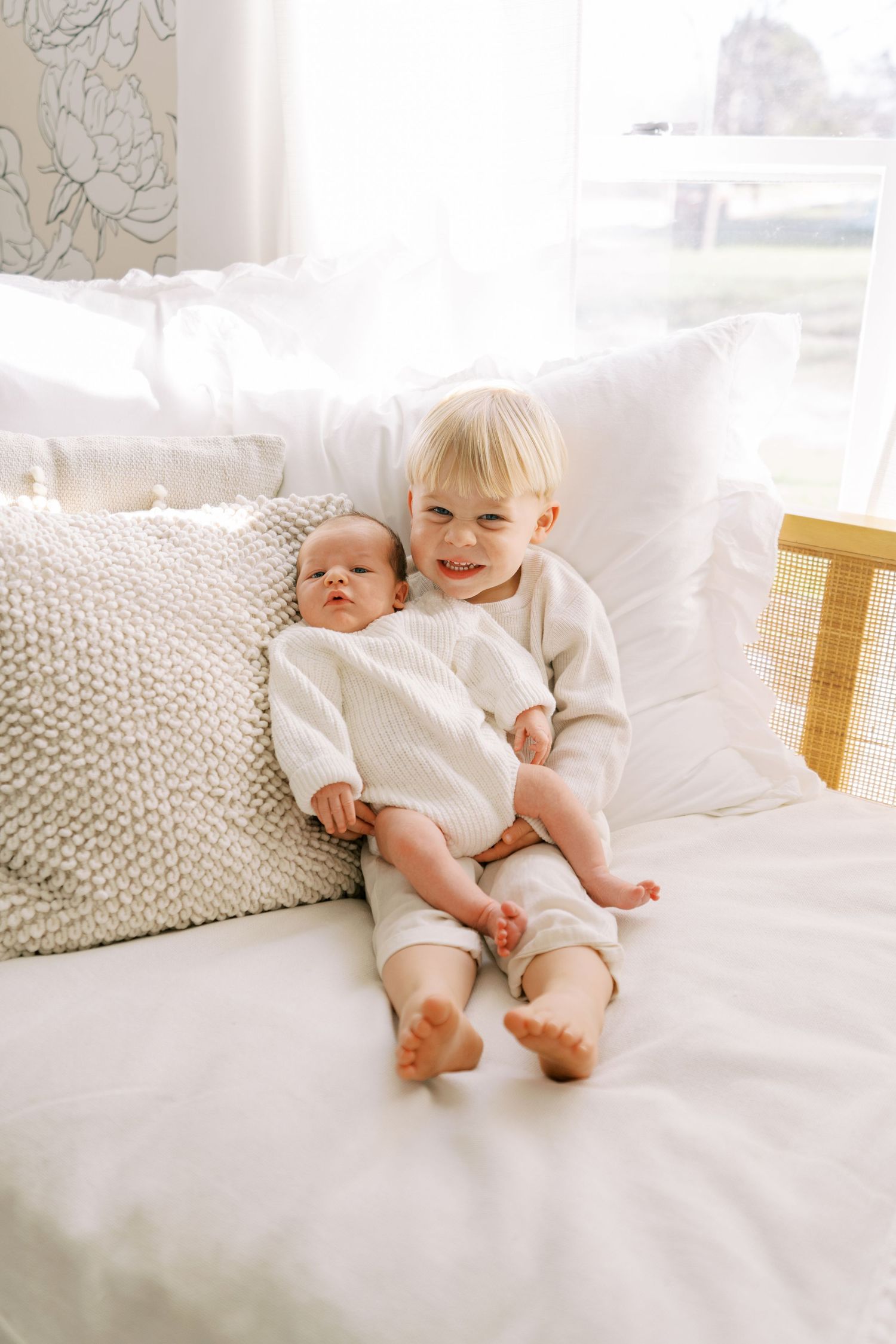 Two young children in white outfits sitting together on a cozy white bed with knit pillows in a bright sunlit room.