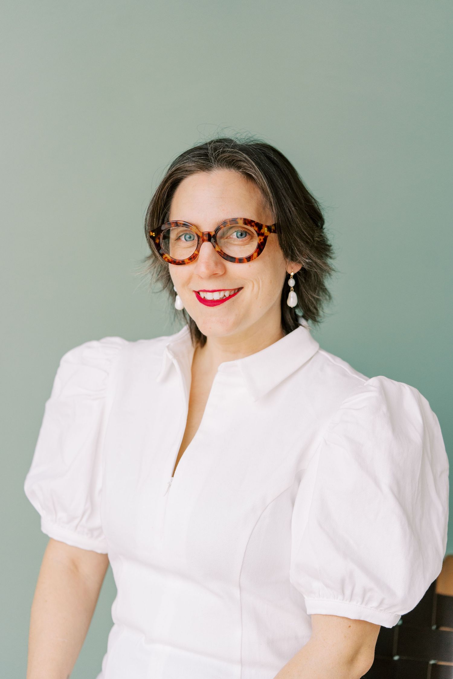 Portrait against sage green wall wearing white dress with statement puff sleeves and vintage-style glasses.
