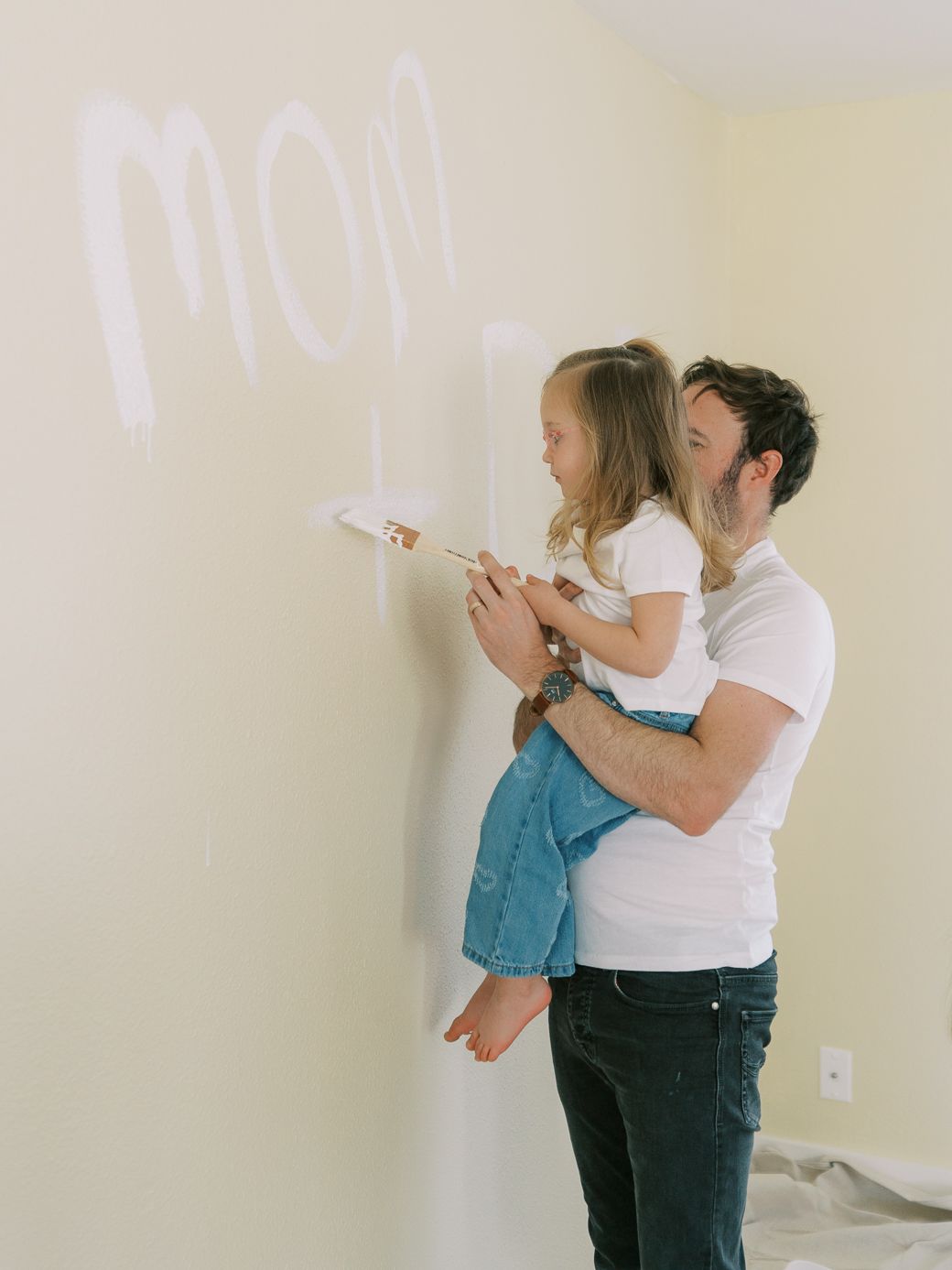 Parent and child write on a wall together while painting a room in soft natural light.