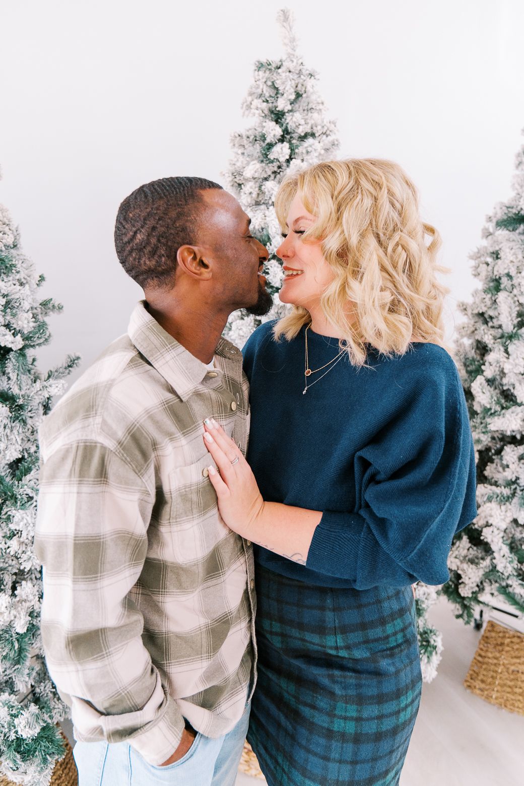 A couple shares a romantic moment in front of decorated white Christmas trees in a bright indoor setting.
