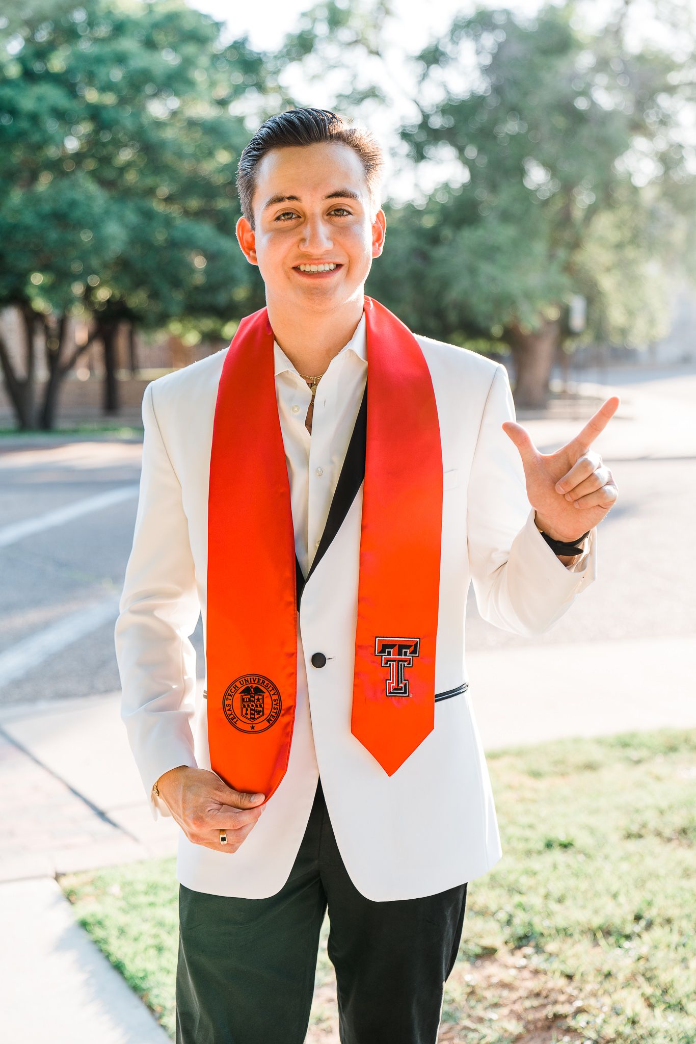 Graduate wearing a white blazer and red stole smiles while posing outdoors for graduation photos on campus.