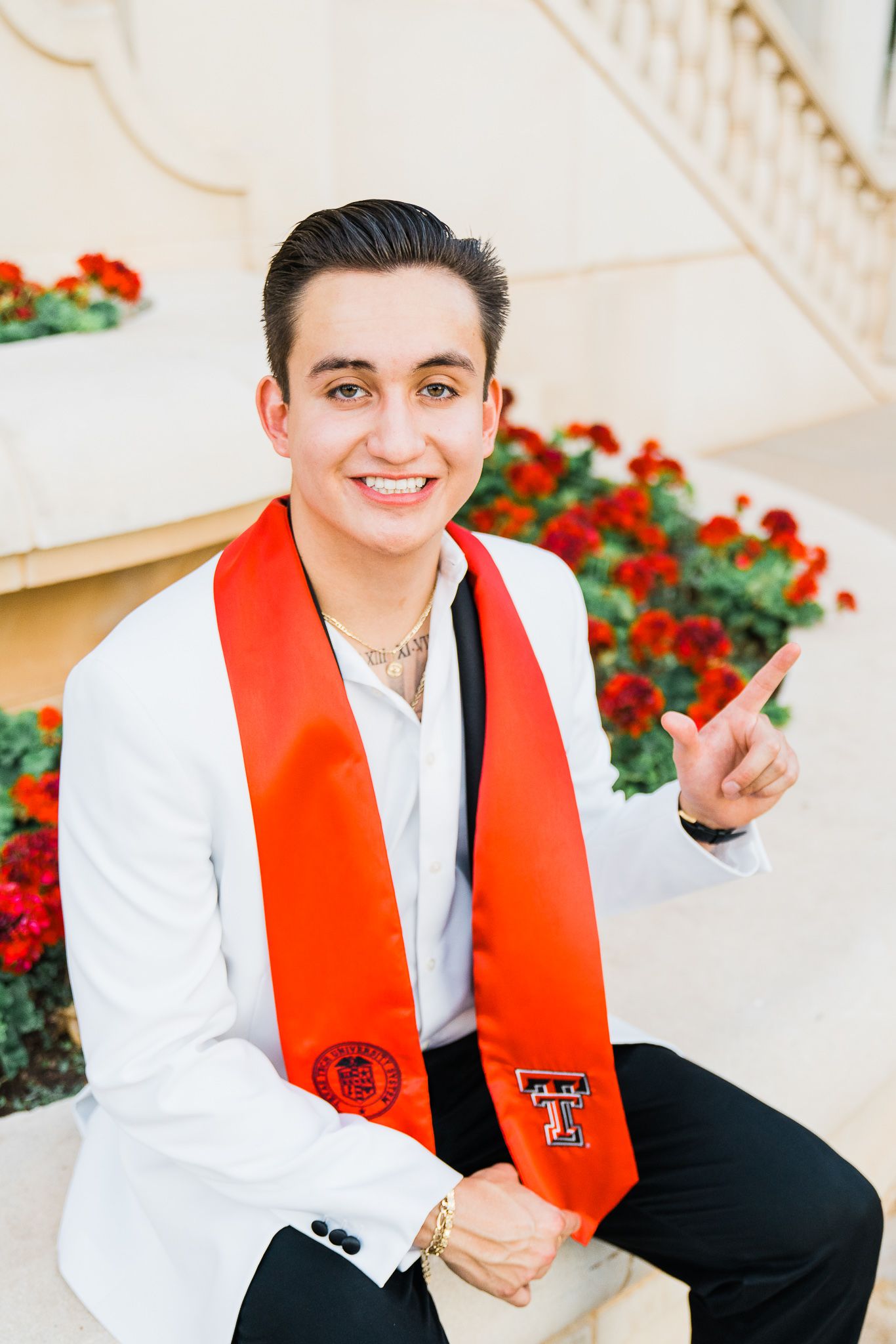 Graduate in white suit and red stole smiling during outdoor photo session with red flowers in background.