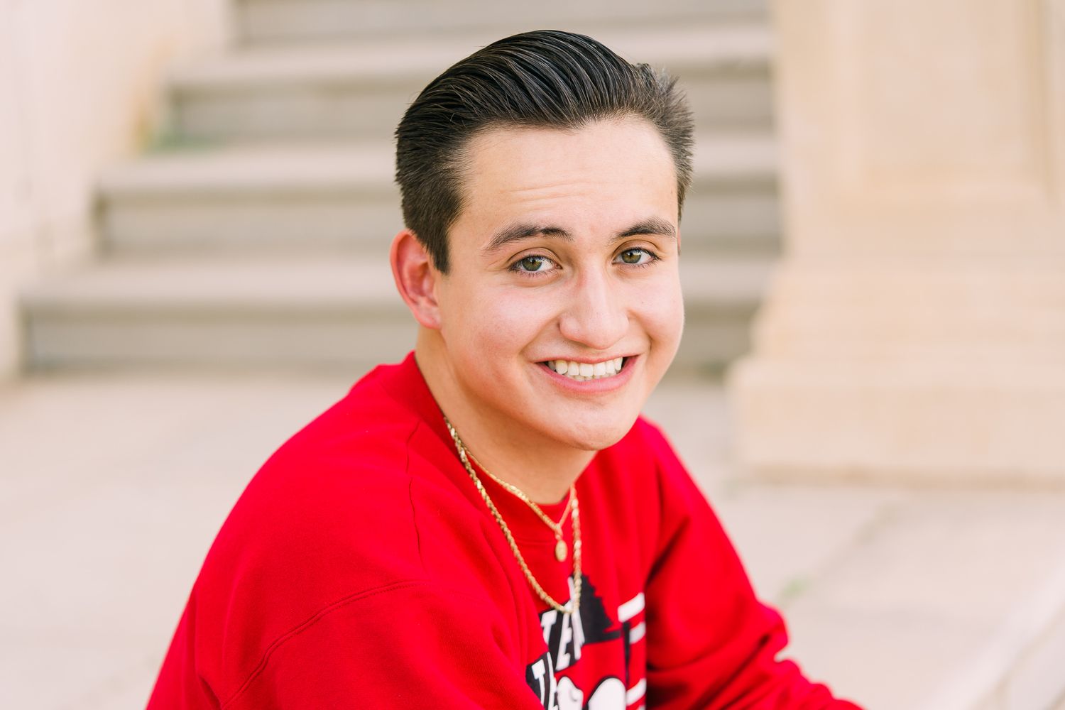 Person in a red sweatshirt smiles while sitting in front of a light colored wall.
