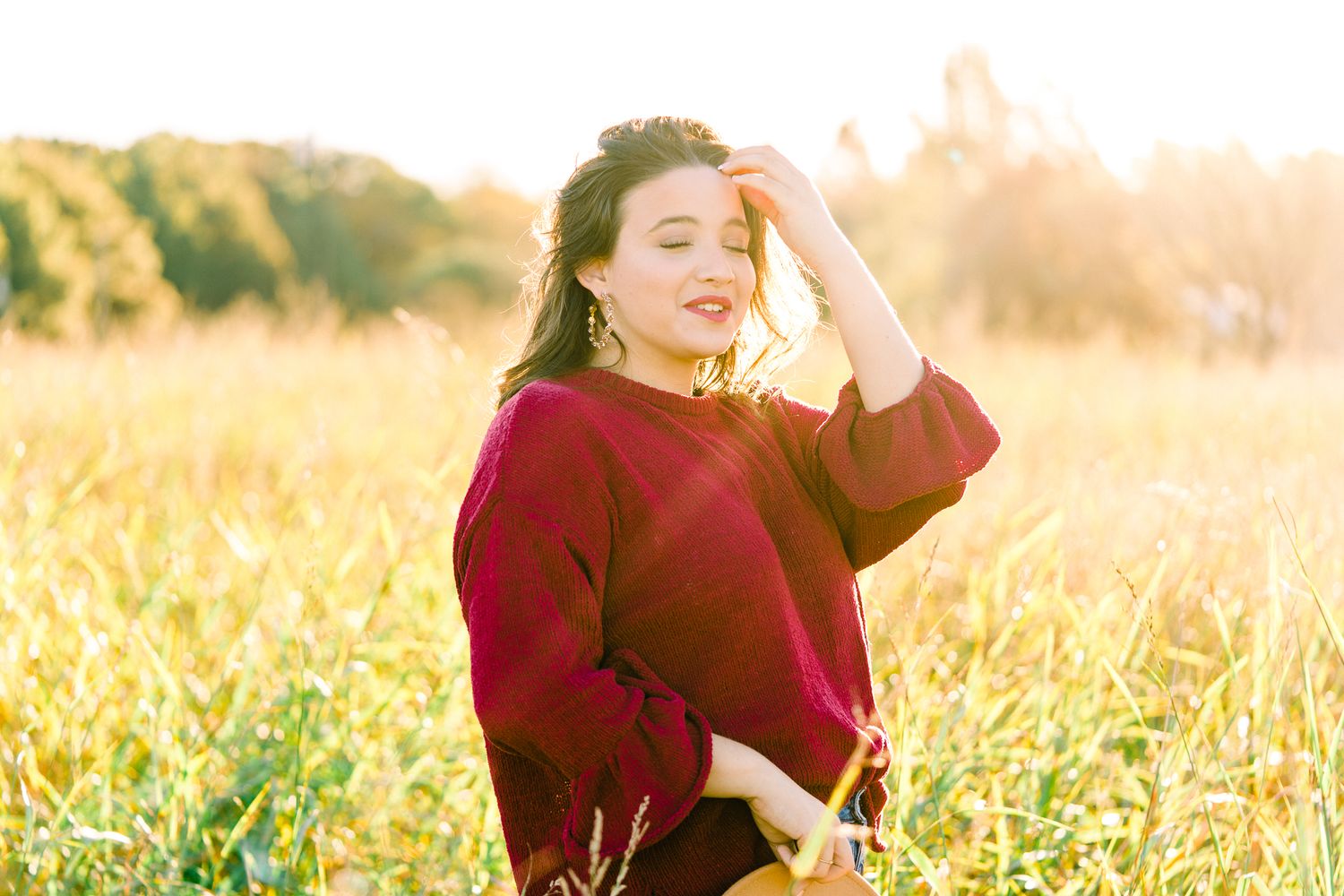Person wearing burgundy sweater stands among tall grass in warm sunlit meadow.