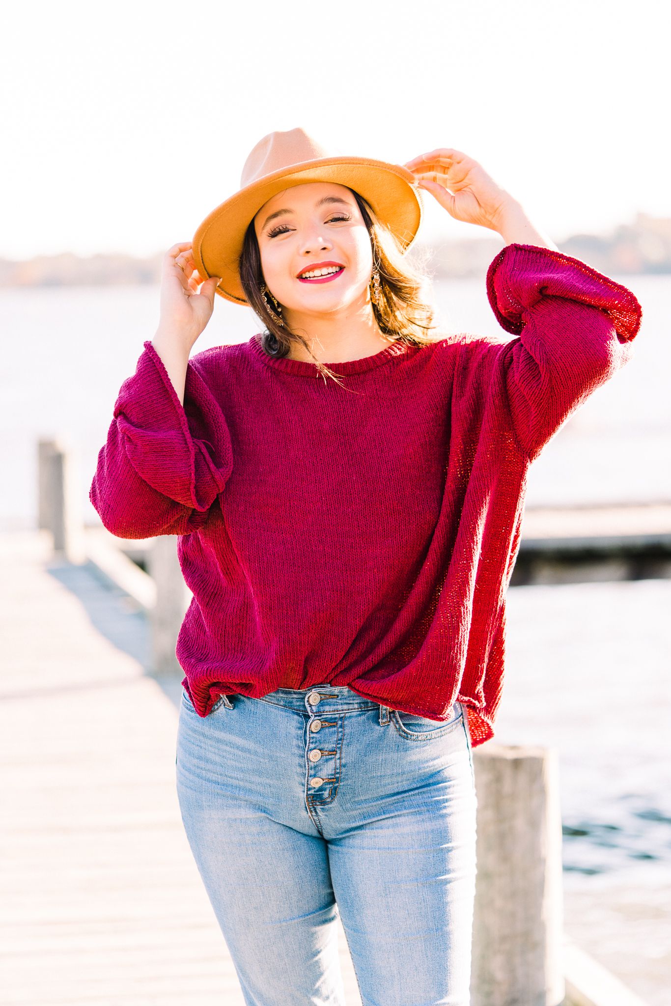 A person in a burgundy sweater and light jeans poses casually outdoors on a sunny day.