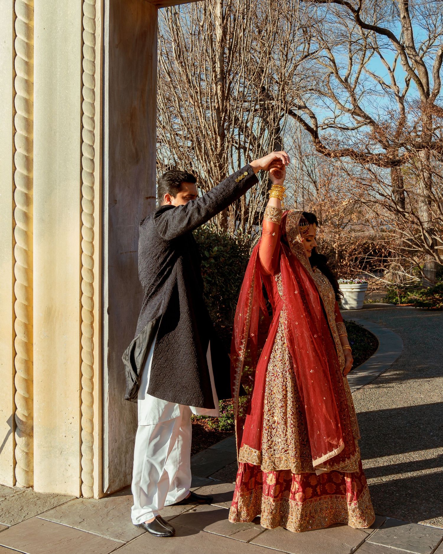 A couple in traditional South Asian wedding attire dancing and posing playfully against architectural columns.