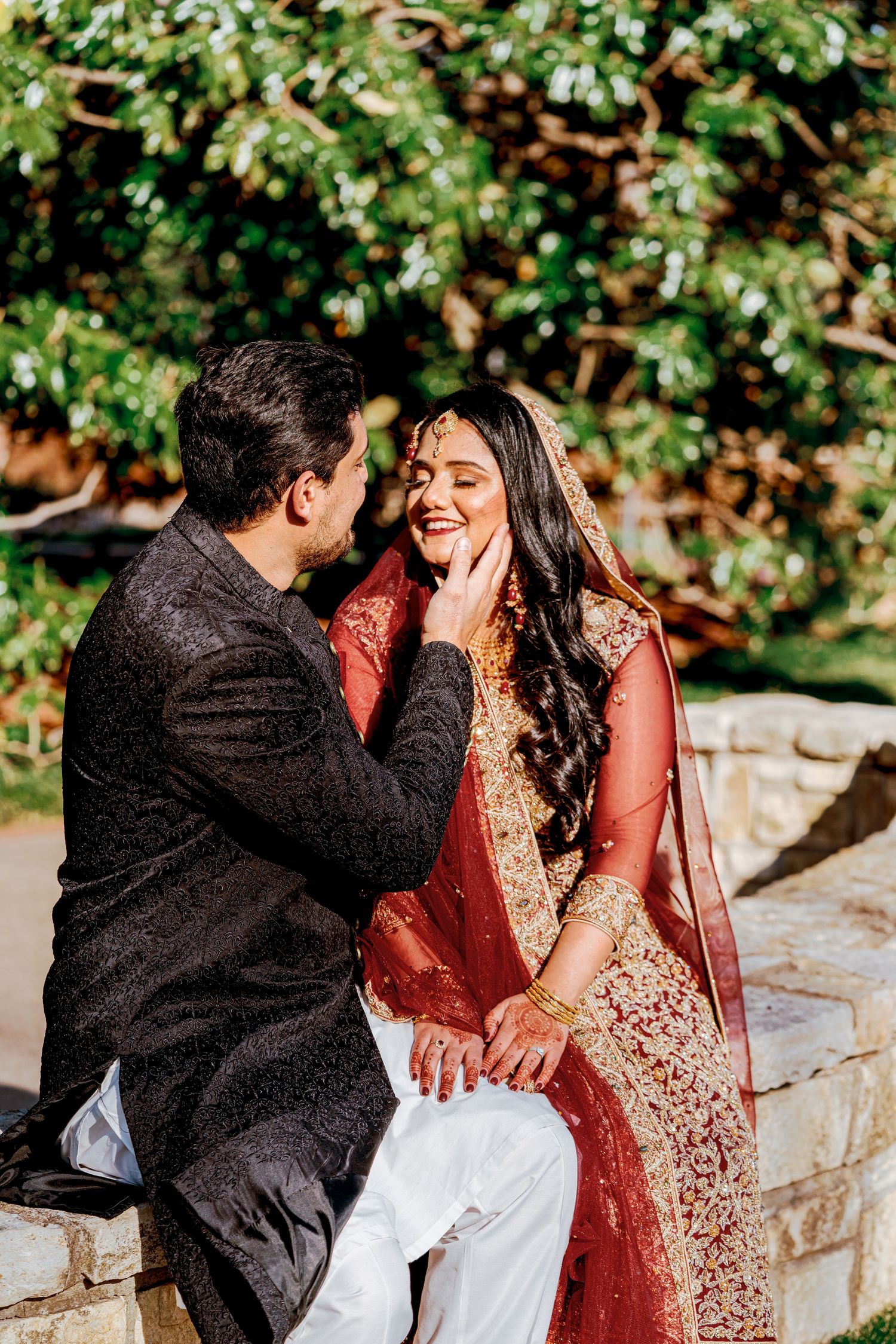 A couple shares a romantic moment in traditional South Asian wedding attire against a garden backdrop.