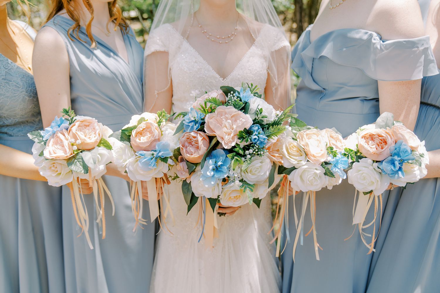 Bridesmaids in dusty blue dresses hold matching peach and blue floral bouquets in a row at an outdoor wedding.