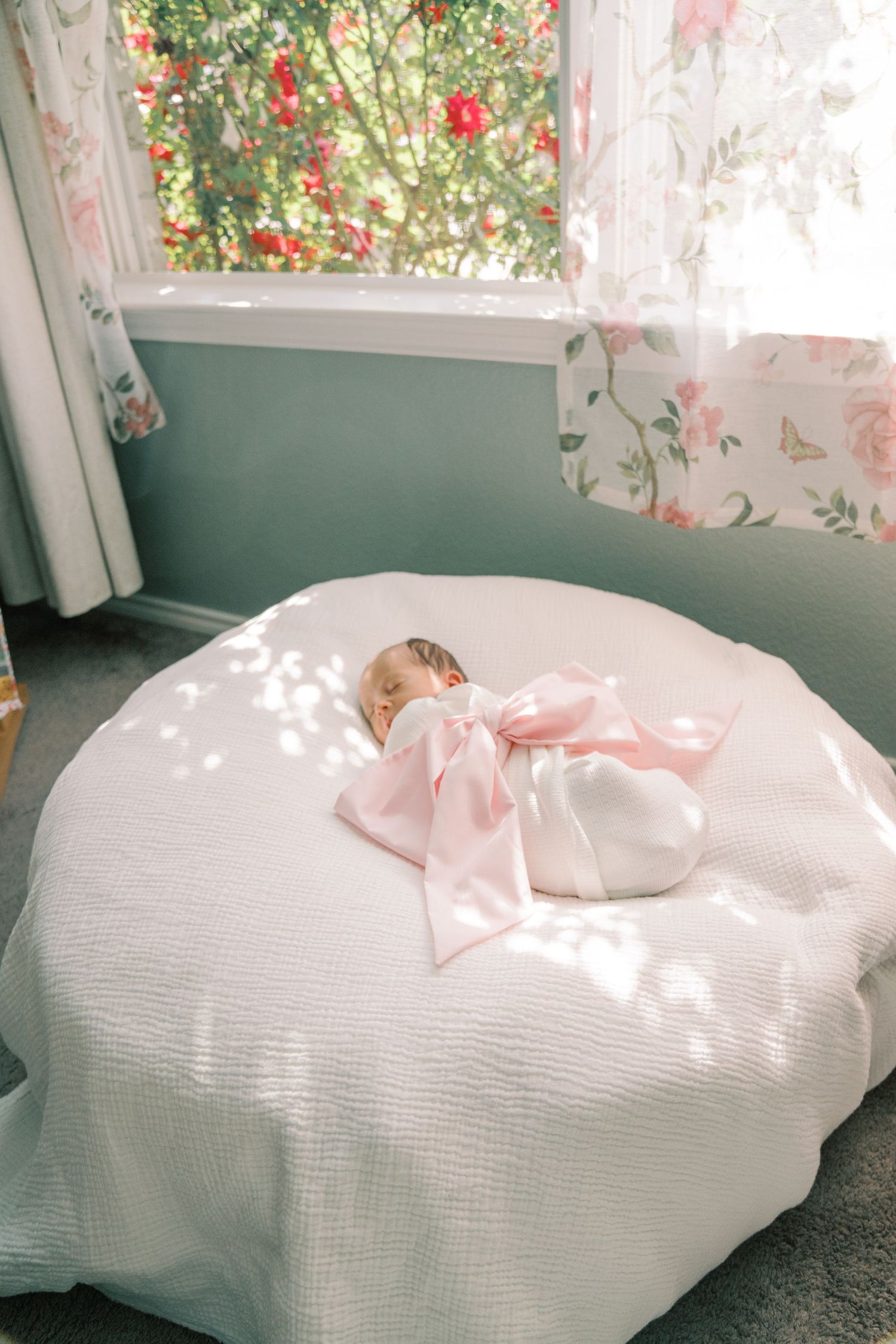 White blanket with pink bow photographed in soft natural light streaming through floral curtains.