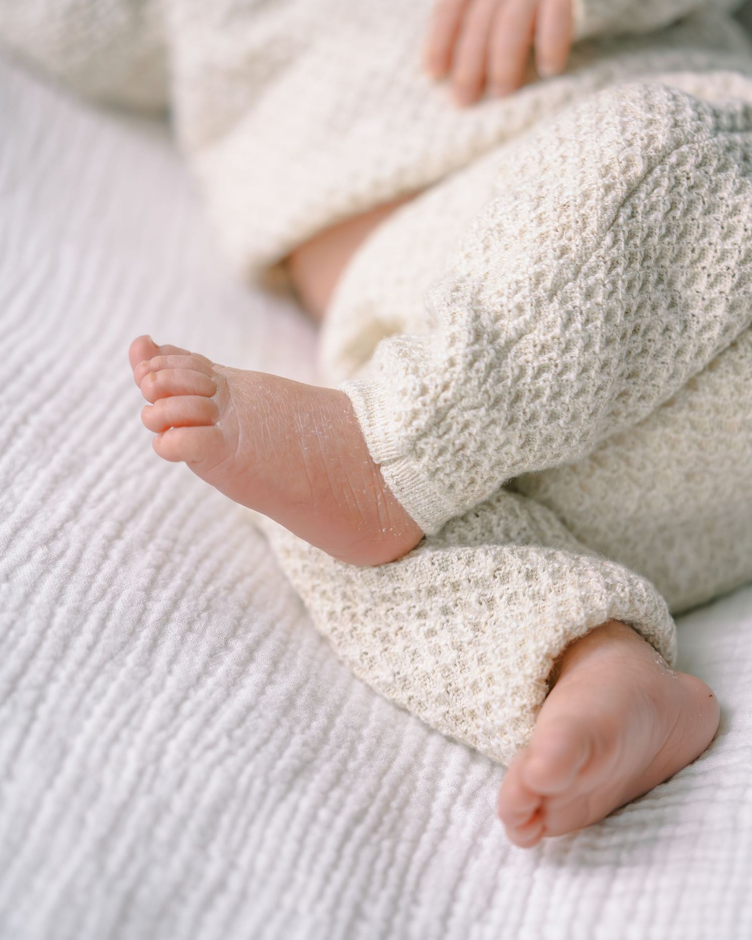 Close-up of a newborn baby's tiny feet and legs in cream knit pants lying on a white textured blanket.