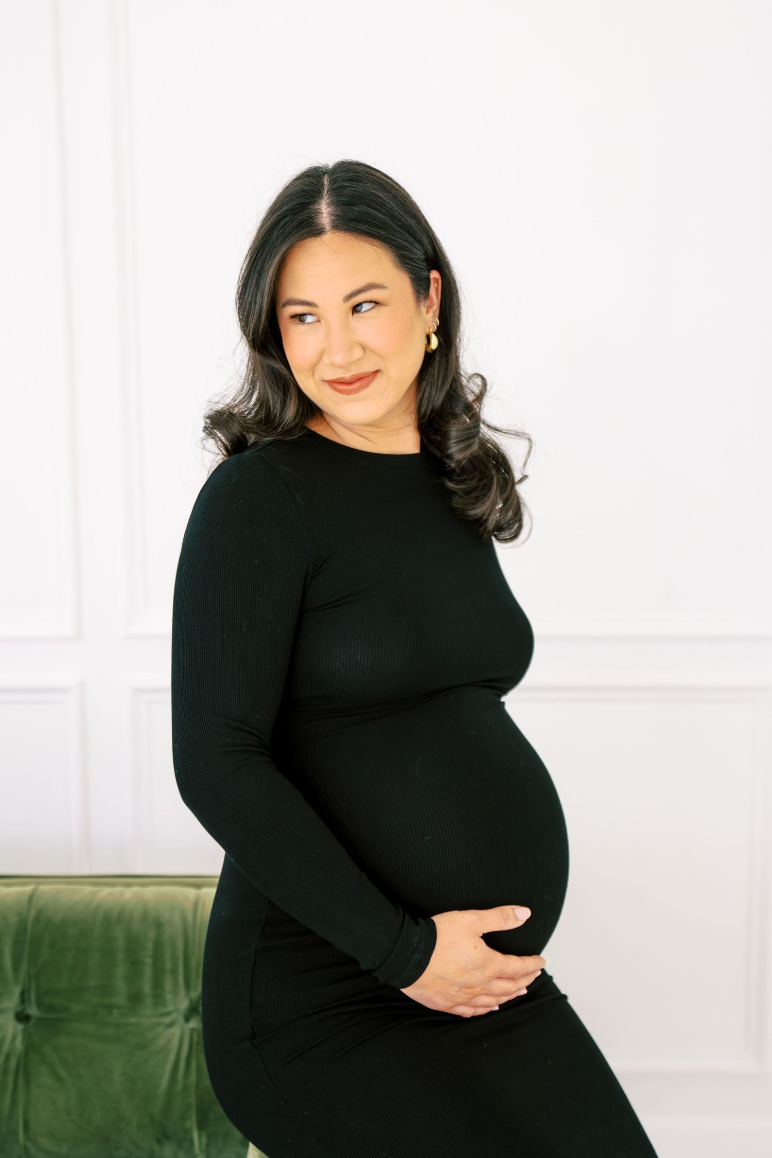 Pregnant woman in black long-sleeve dress cradling baby bump, smiling softly in bright white studio setting.