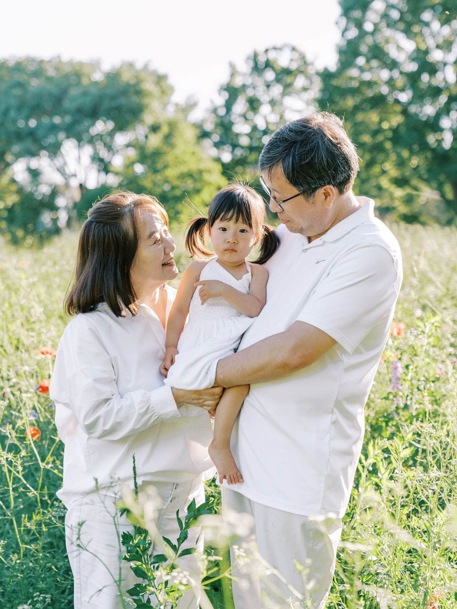 Family of three in white outfits posing in a sunny green meadow with wildflowers, parents smiling at toddler daughter.