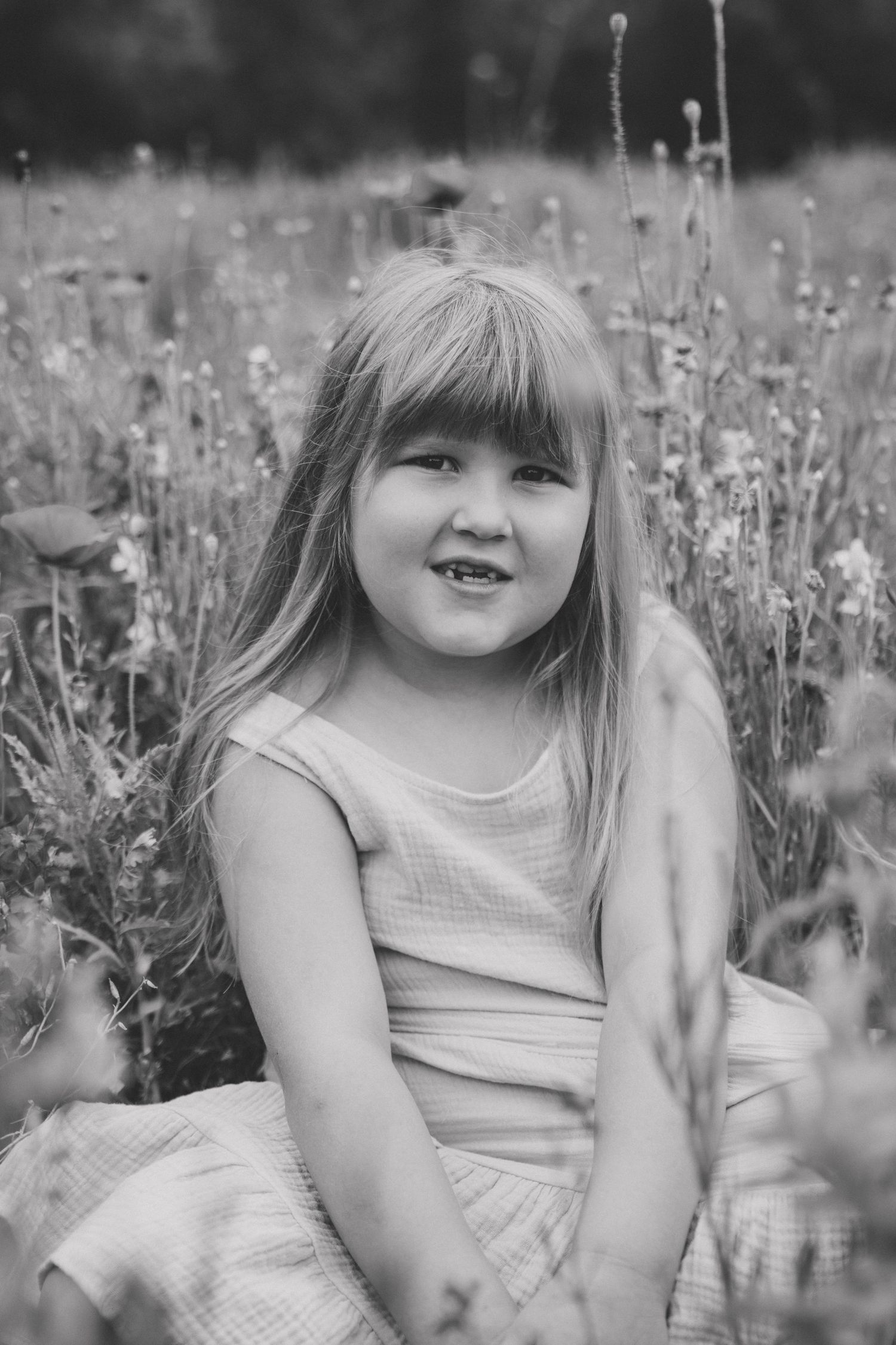 Black and white portrait of a smiling young girl with long hair sitting in a wildflower meadow wearing a sleeveless dress.