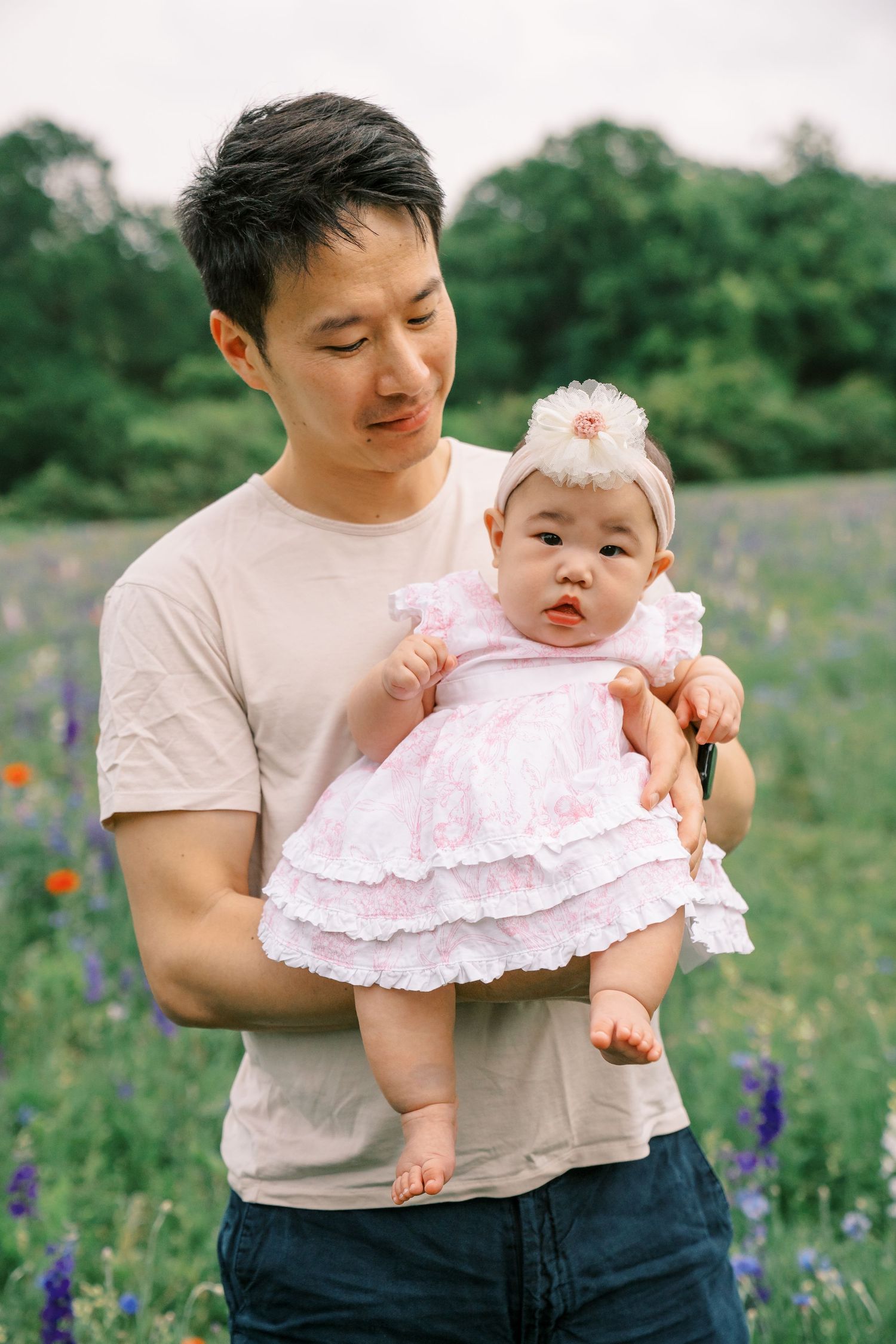 Father holding baby girl in pink dress in flower field.