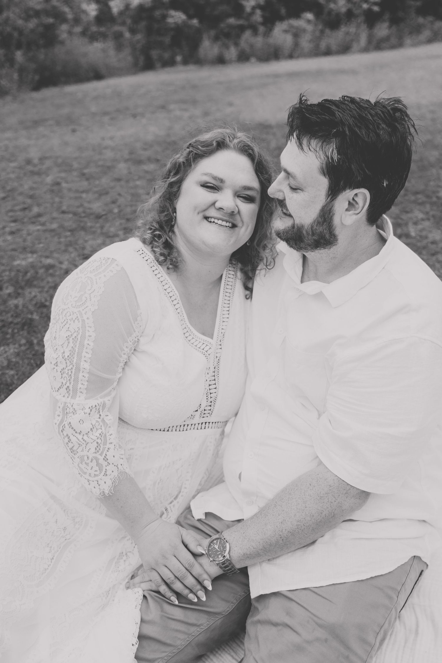 Black and white photo of smiling couple sitting together outdoors on a blanket in a park setting.