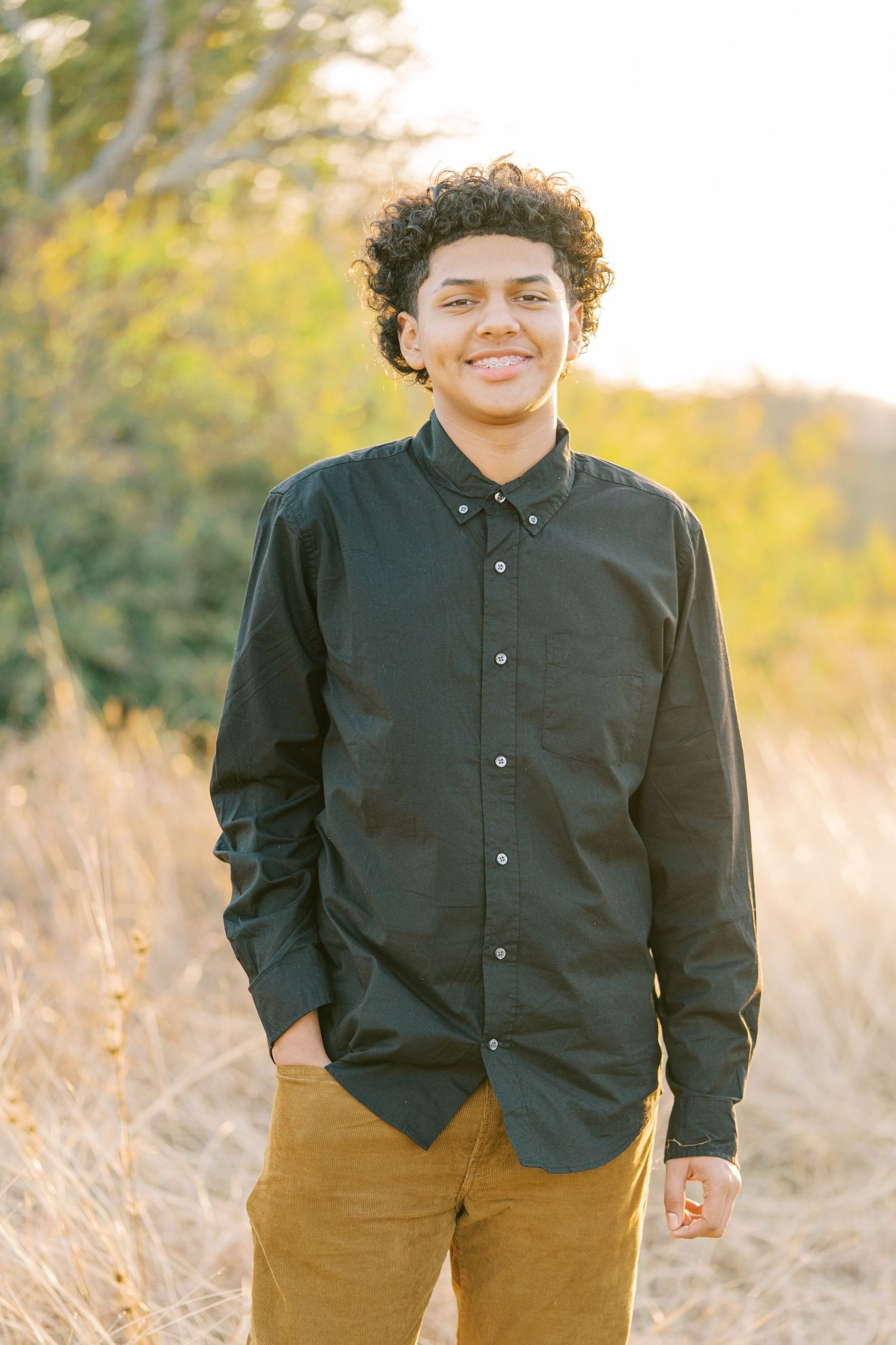 A person in a black button-down shirt and brown pants smiles warmly while standing in a sunlit field.