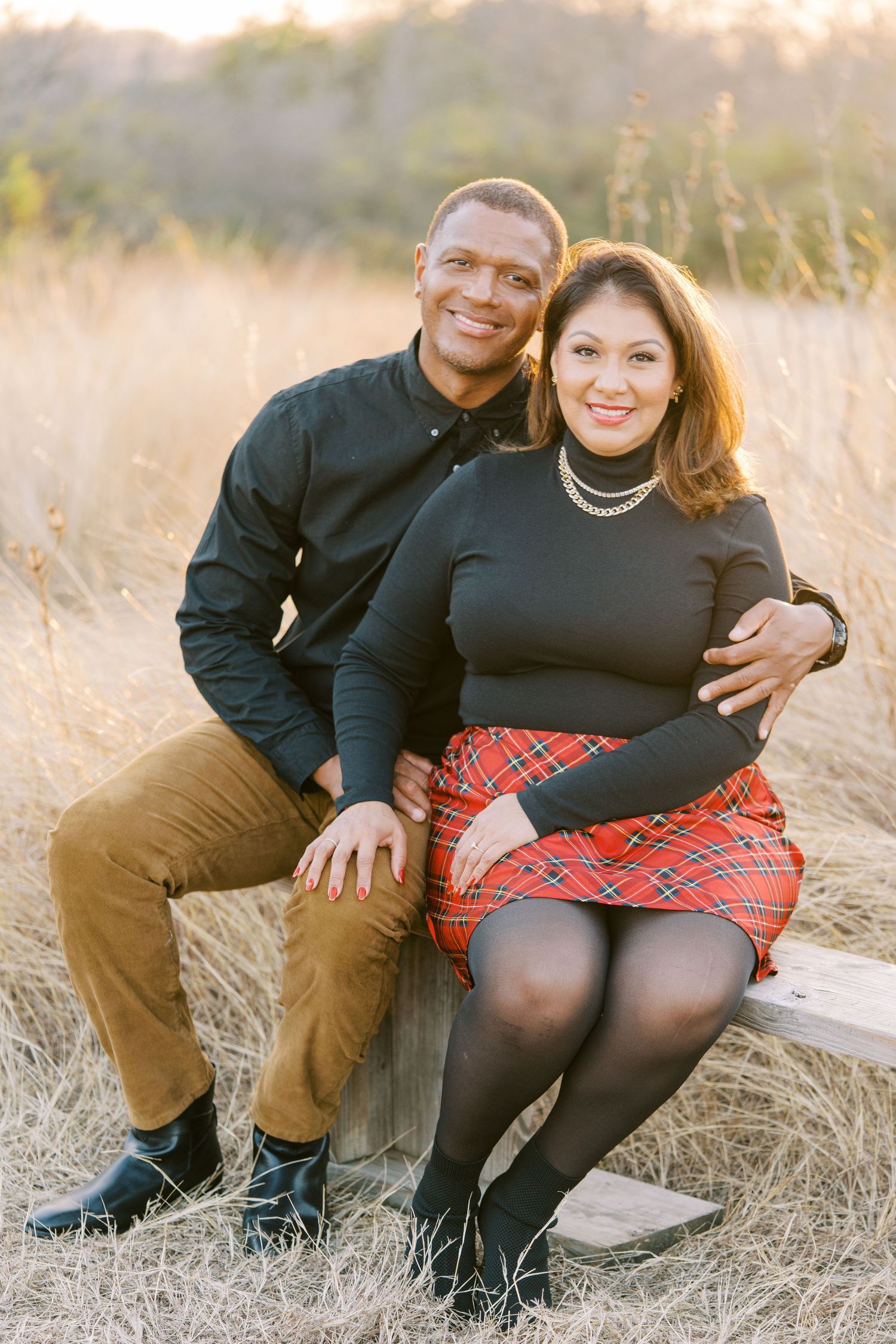 A couple sits together on rustic wooden steps surrounded by tall golden grass at sunset.