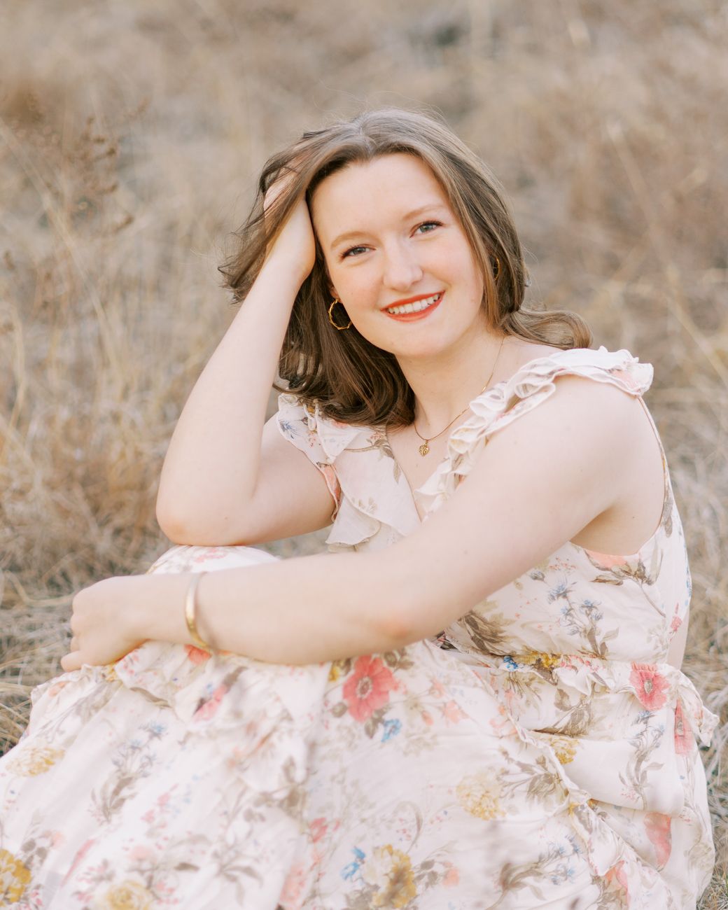A person in a floral dress poses gracefully in a field of dried grass during a golden hour photoshoot.