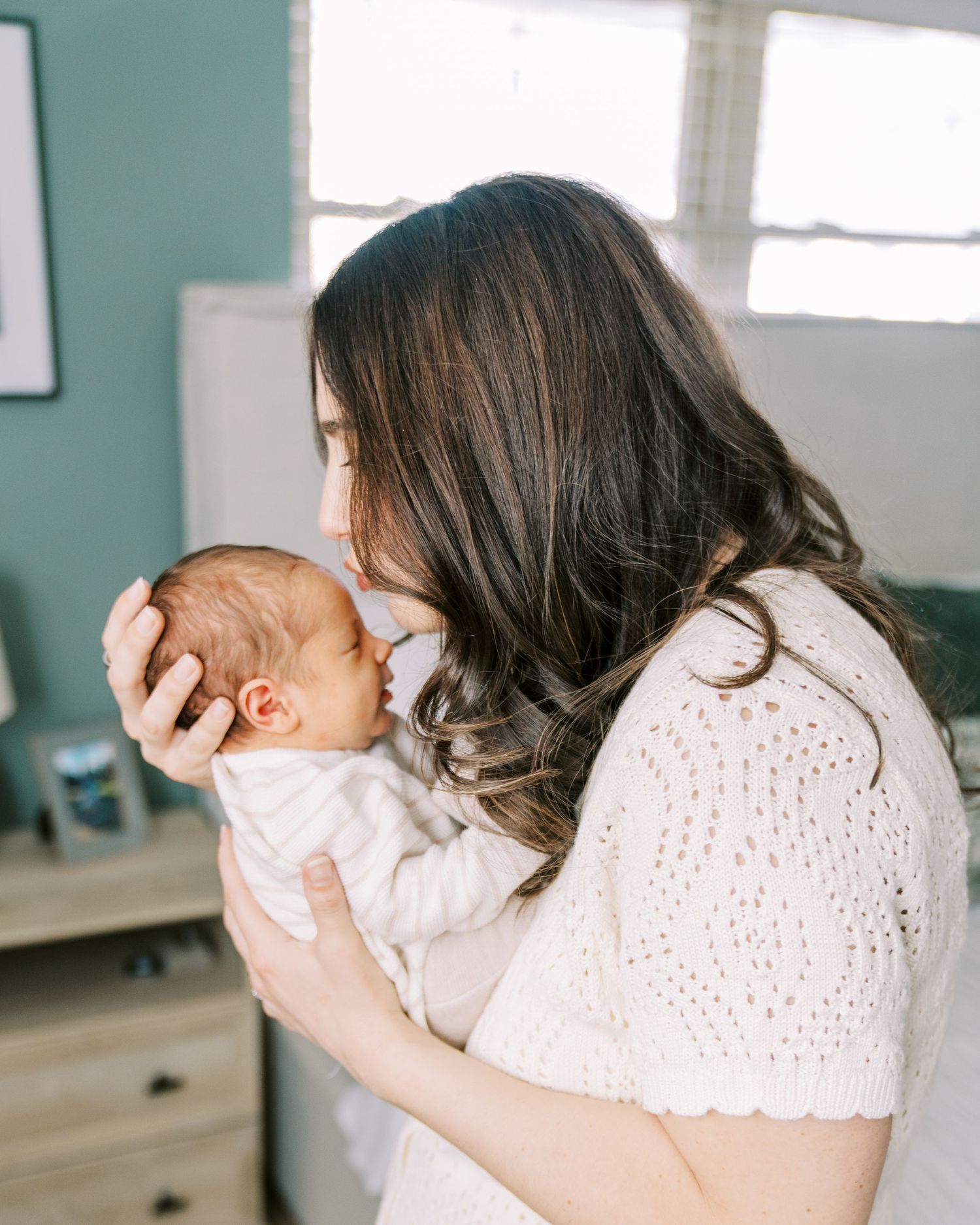 Mother in white eyelet dress holding and kissing her newborn baby in a bright studio setting.