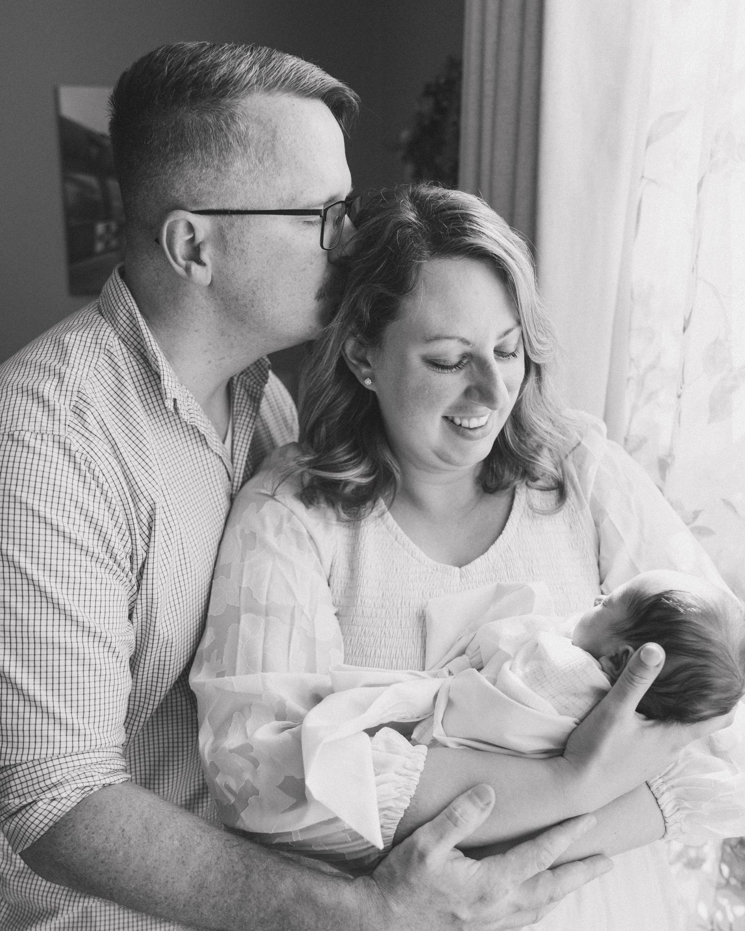 A tender black and white portrait of a couple with their newborn baby by a window, sharing an intimate family moment.