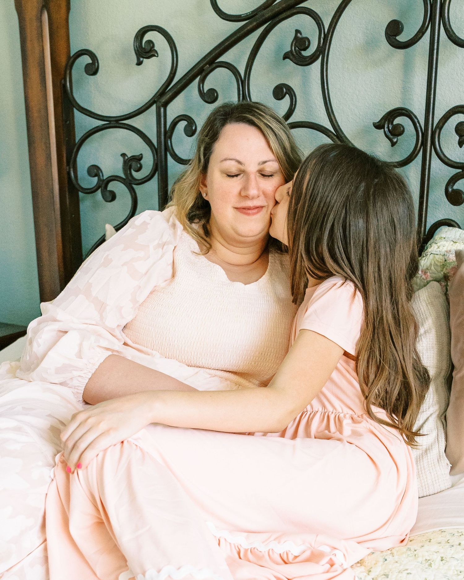 Family shares tender moments together on bed with ornate metal headboard in soft pink tones.