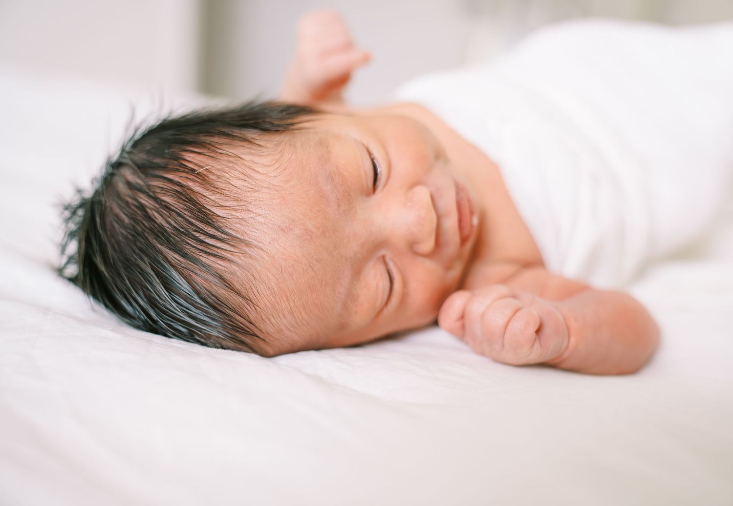 Peaceful newborn baby with dark hair sleeping on white bedding, eyes closed with a gentle smile.