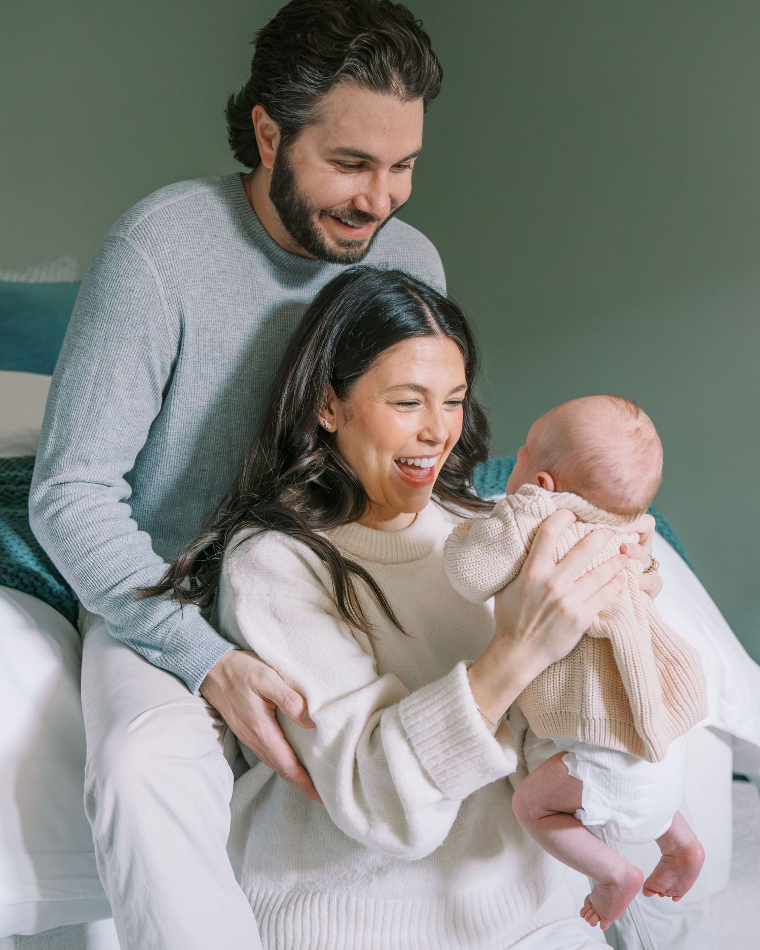 Happy couple smiling at their newborn baby, mother in white sweater holding infant, father in gray sweater, indoor setting.