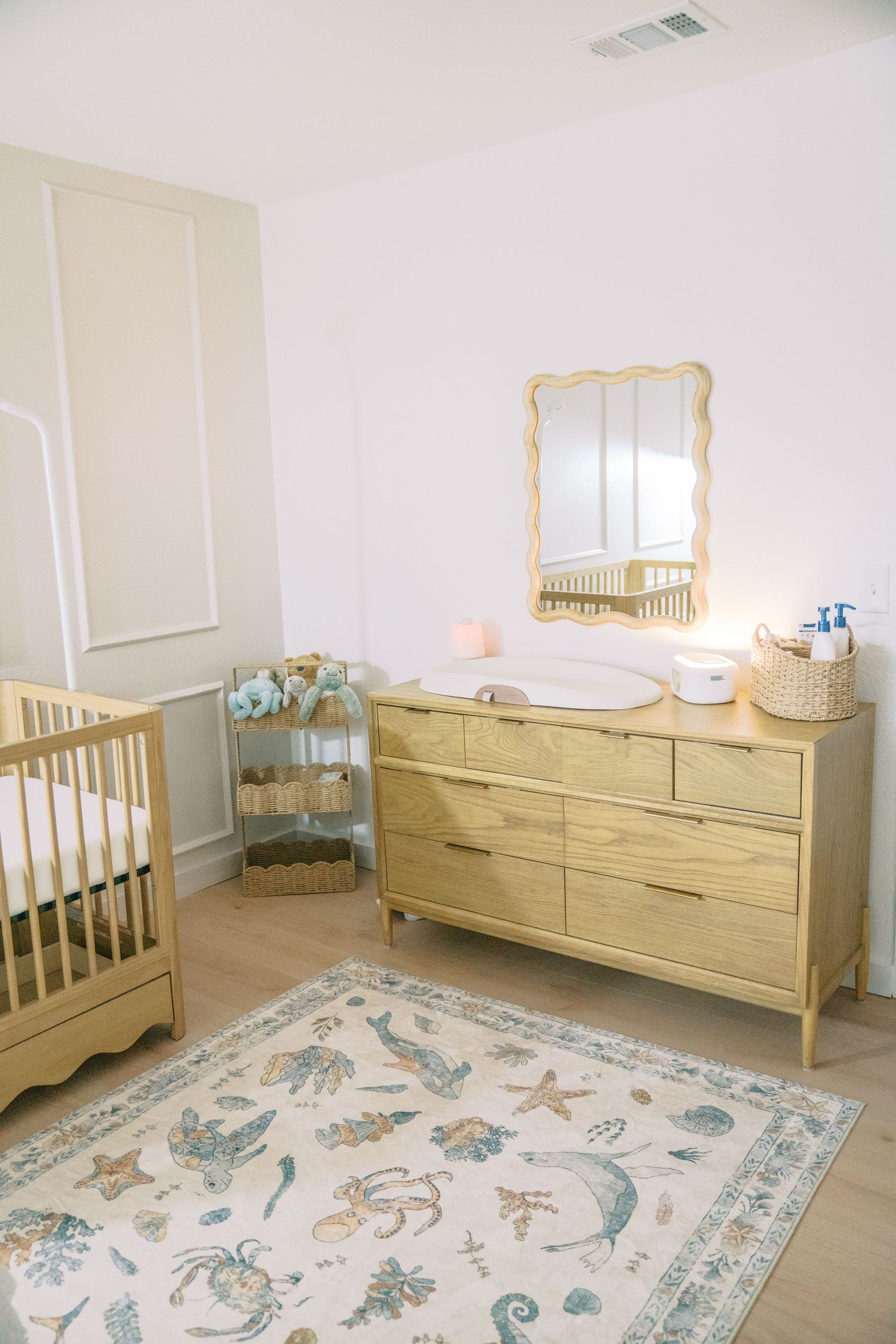 Bright nursery with wooden crib, dresser, wavy gold mirror, ocean-themed rug, and soft neutral decor.
