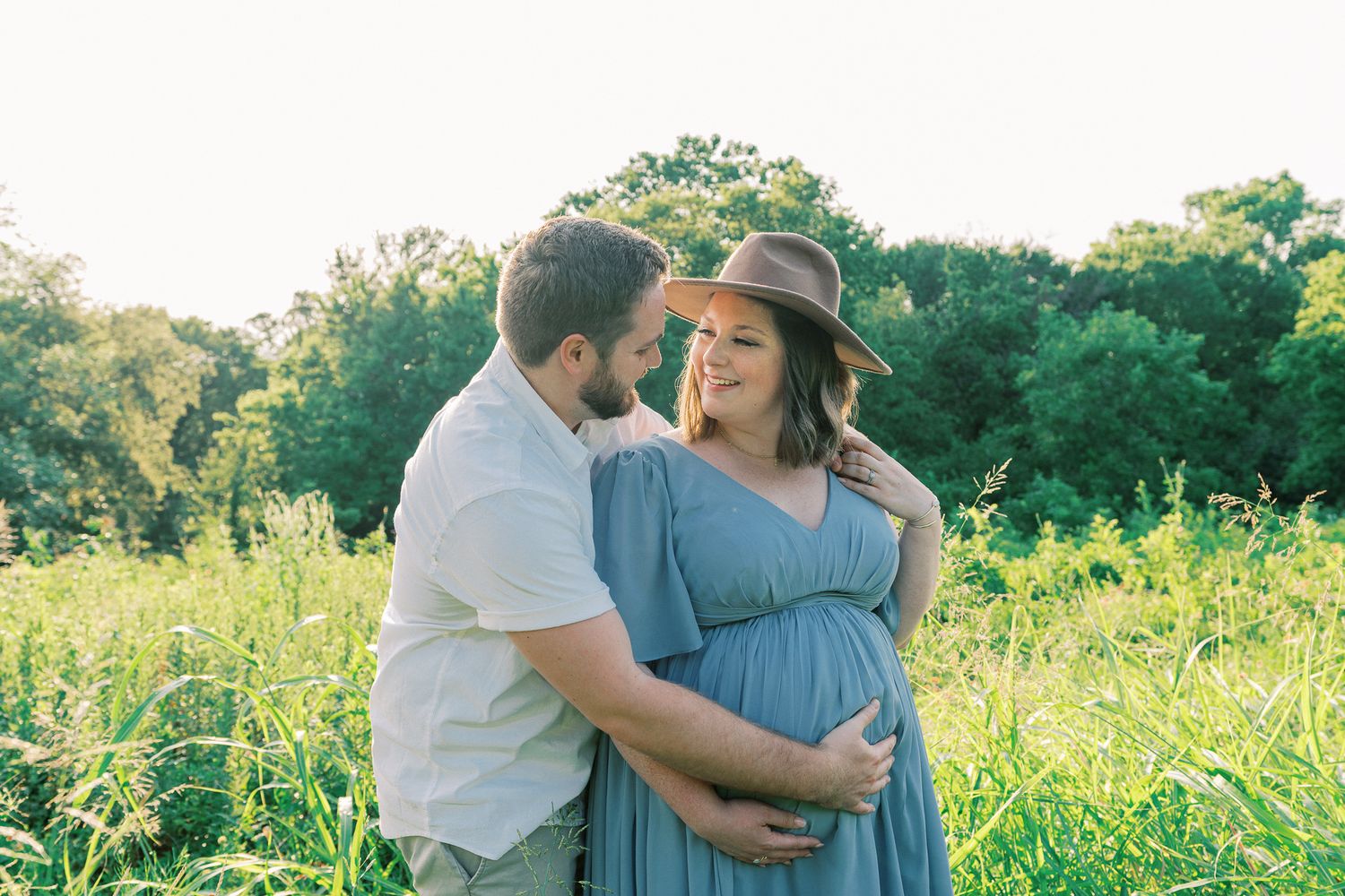 A couple embraces in a sunny meadow with wildflowers, the woman wearing a blue dress and brown hat.
