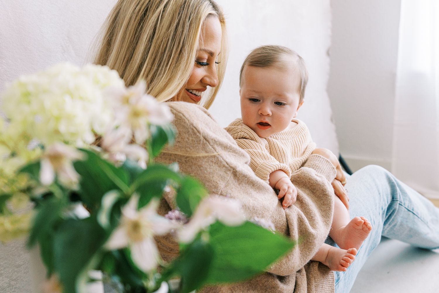 A sweet indoor portrait featuring a parent holding a baby with soft natural light and floral decor.